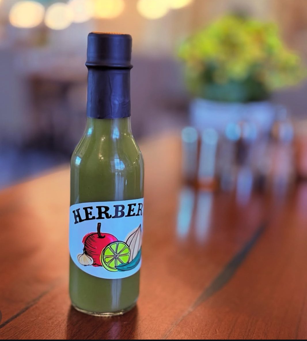 A small green bottle with a black cap and a colorful label that says "Herber" with illustrations of garlic, lime slices, and a chili pepper on a wooden table. Out of focus potted plant in the background.
