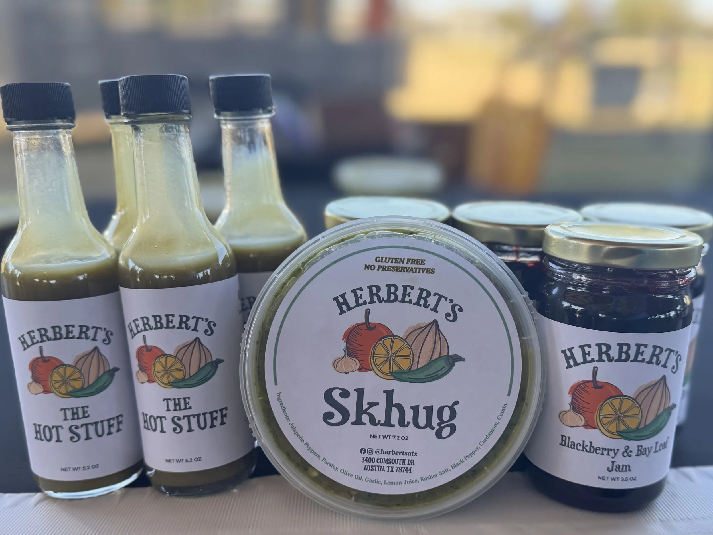 Collection of condiment jars and bottles on a table, including three small glass bottles labeled 'Herbert's The Hot Stuff', a container labeled 'Herbert's Skhug', and a jar labeled 'Blackberry & Bay Leaf Jam'.