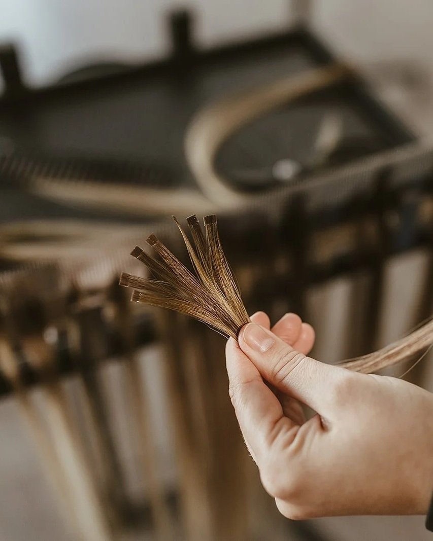 A hand holds a small bundle of hair extension strands in front of a blurred background, possibly in a salon or hair extension shop.