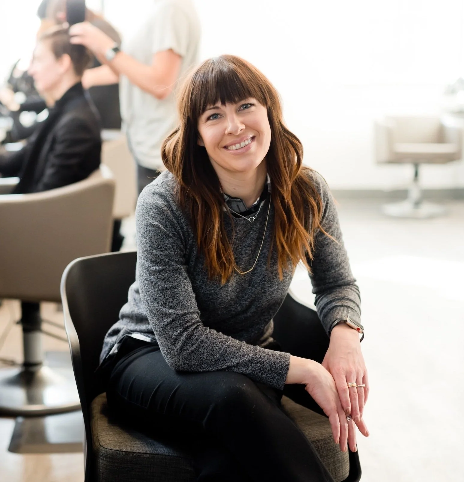 A woman with brown hair and bangs sitting on a chair, smiling at the camera, in a busy salon or beauty parlor.