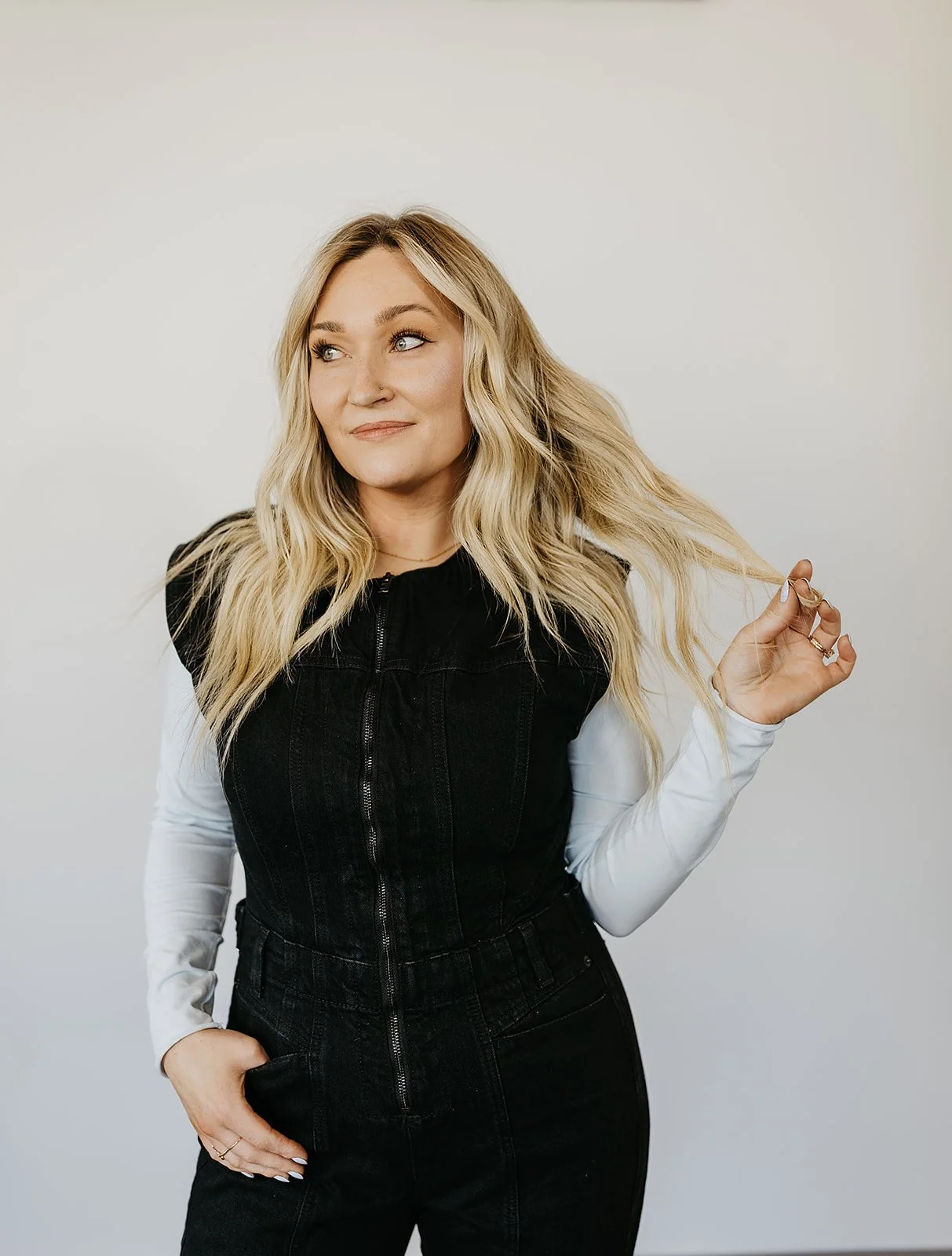 A woman with long, wavy blonde hair, wearing a black sleeveless jacket over a white long-sleeve shirt, poses against a plain light background.