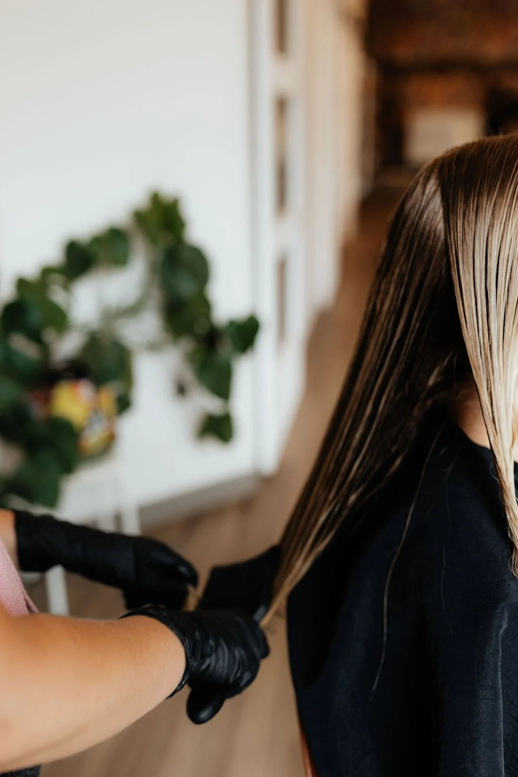 Person with blonde hair getting hair dyed, hairstylist wearing black gloves applying dye to hair in a salon.