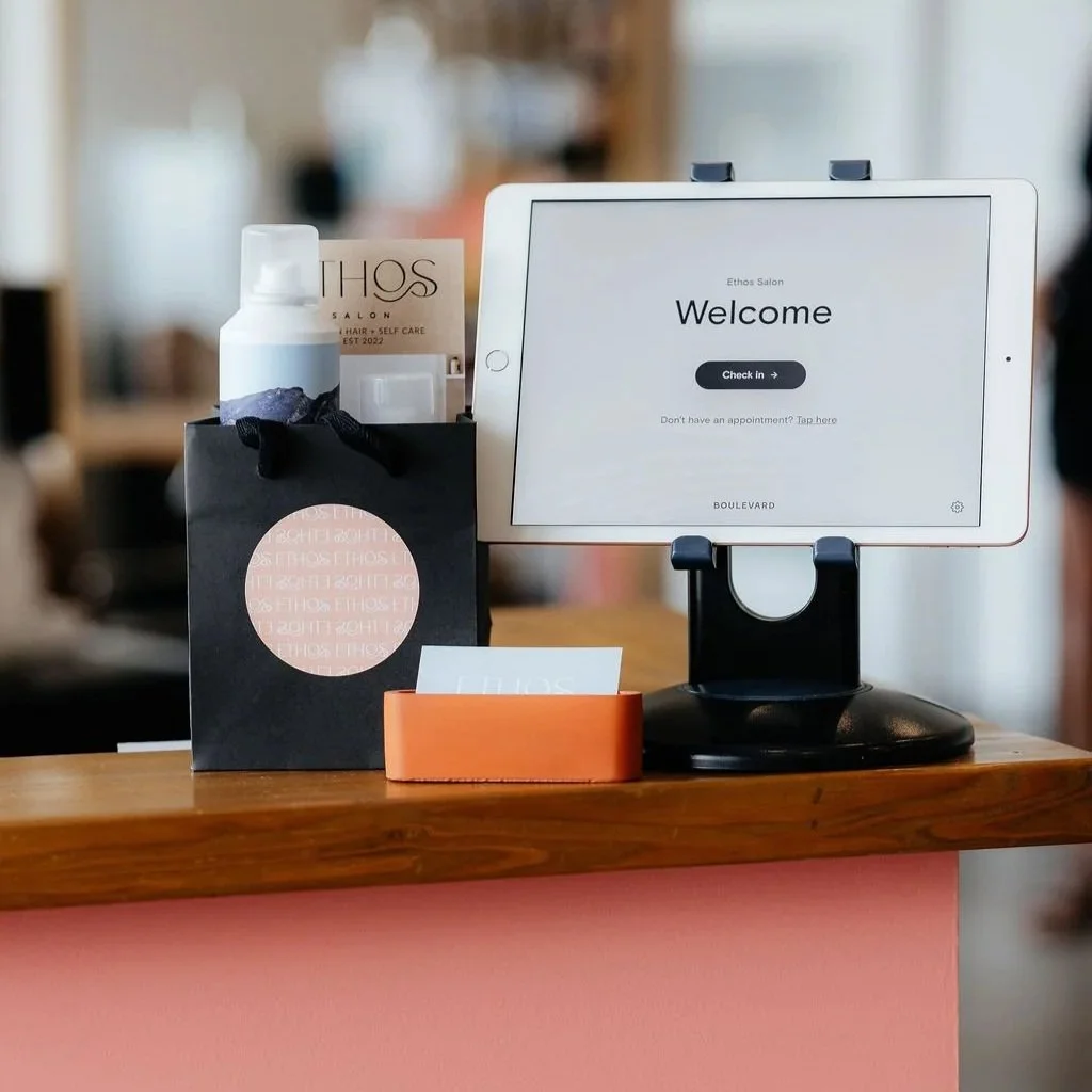 Reception desk at Ethos Salon with check-in tablet, black gift bag, bottle of product, and orange note holder.