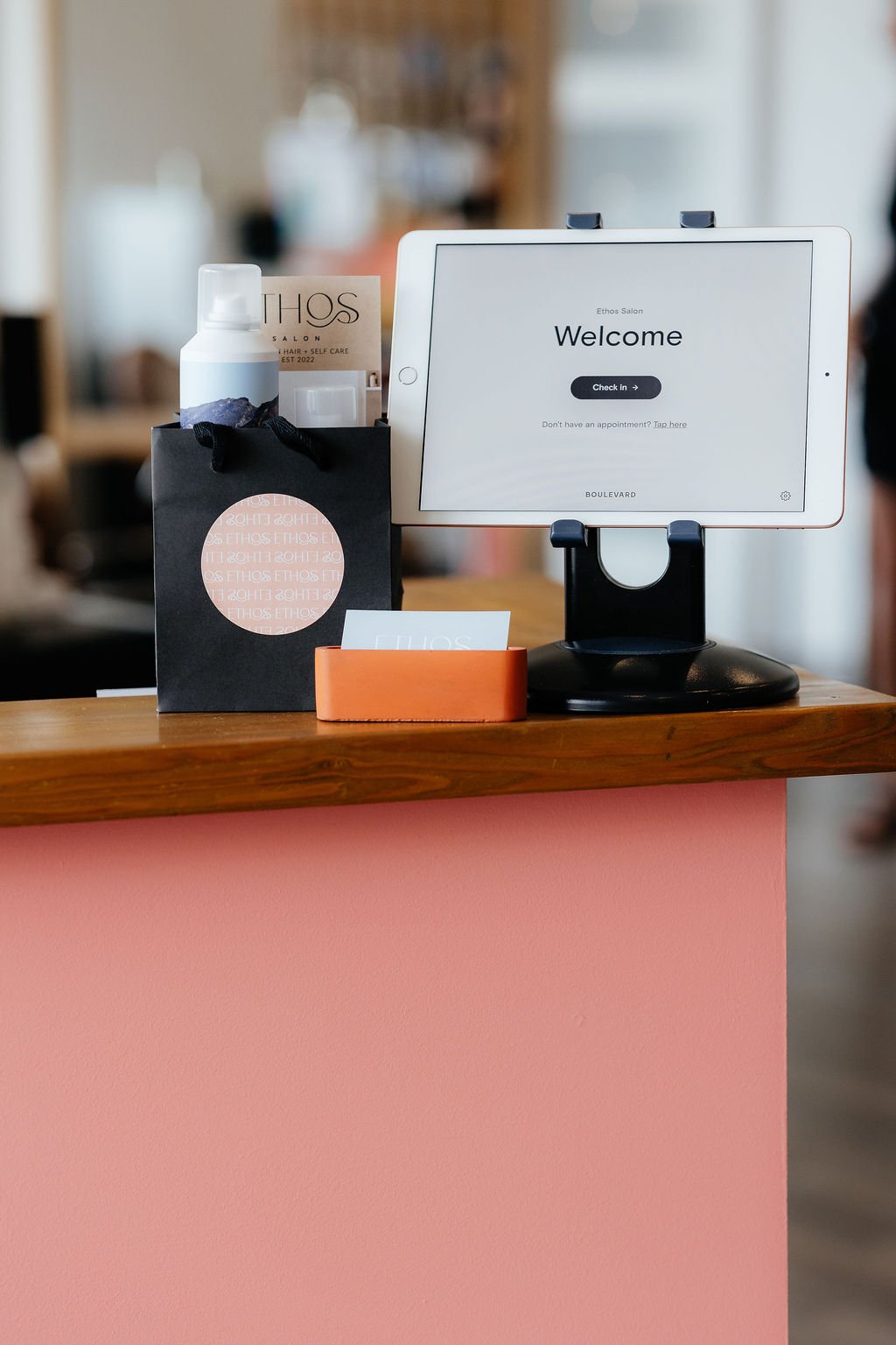 Reception desk with a tablet displaying a welcome screen, a black gift bag with a business card, and a small orange container on a pink countertop at Ethos Salon.