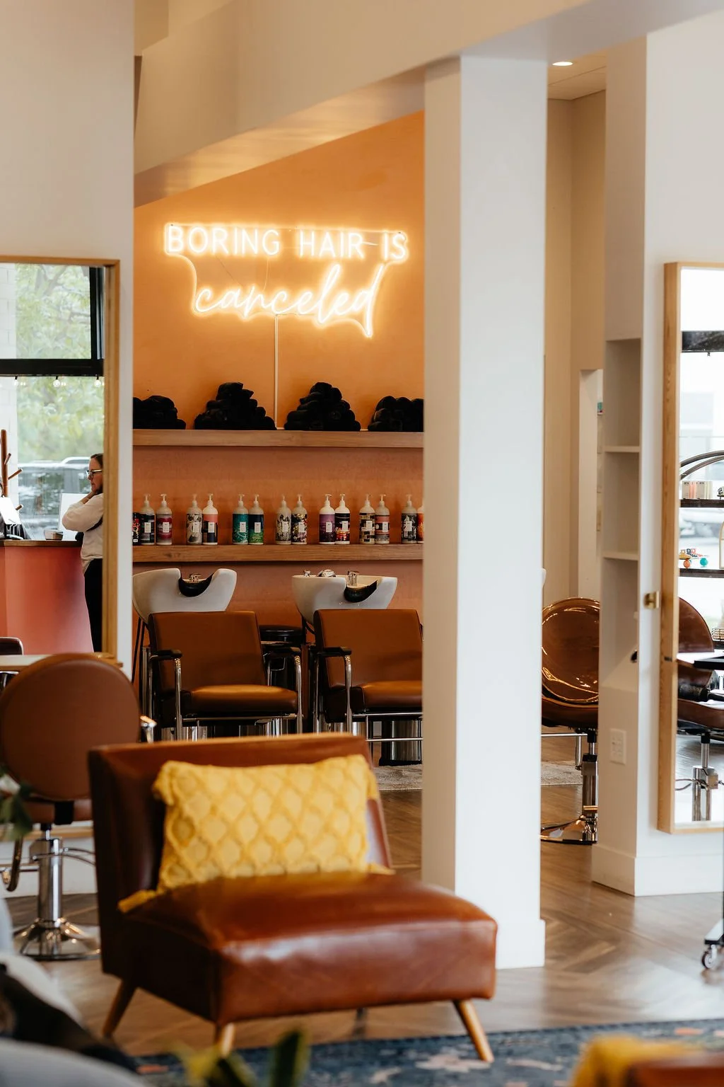 Interior of a hair salon with a neon sign that reads 'BORING HAIR IS canceled,' shampoo stations, chairs, and bottles of hair products on shelves.