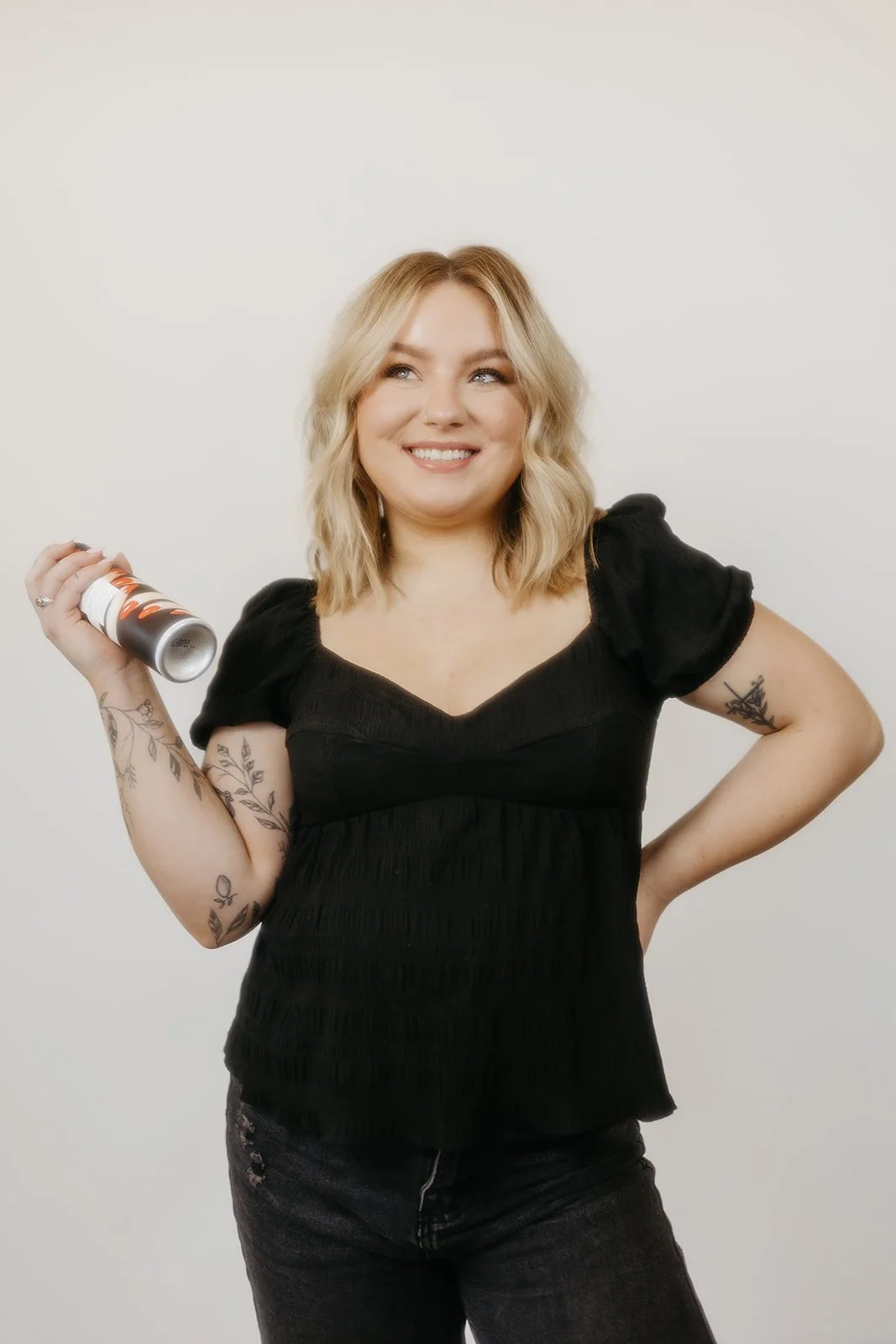 A woman with blonde hair in loose waves, wearing a black short-sleeve top and dark jeans, smiling and holding a can of spray paint in her right hand, standing against a plain white background.