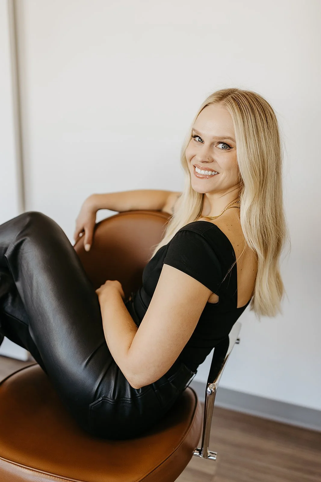 A woman with blonde hair and a nose piercing smiling while sitting sideways on a brown chair, wearing a black top and leather pants.
