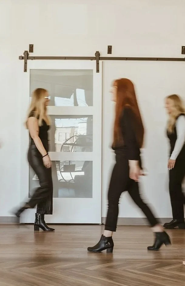 Three women walking past a white wall with a sliding barn door in an indoor space.