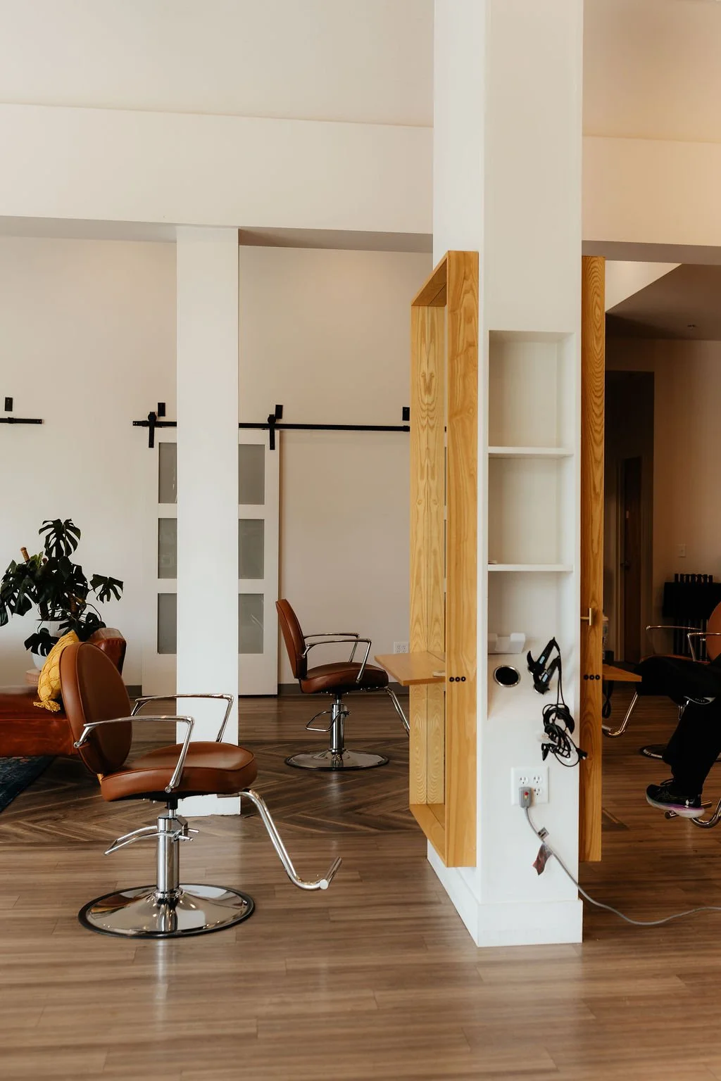 A modern interior with brown salon chairs, a potted plant, white sliding barn doors, and a wooden partition with shelves and headphones hanging.