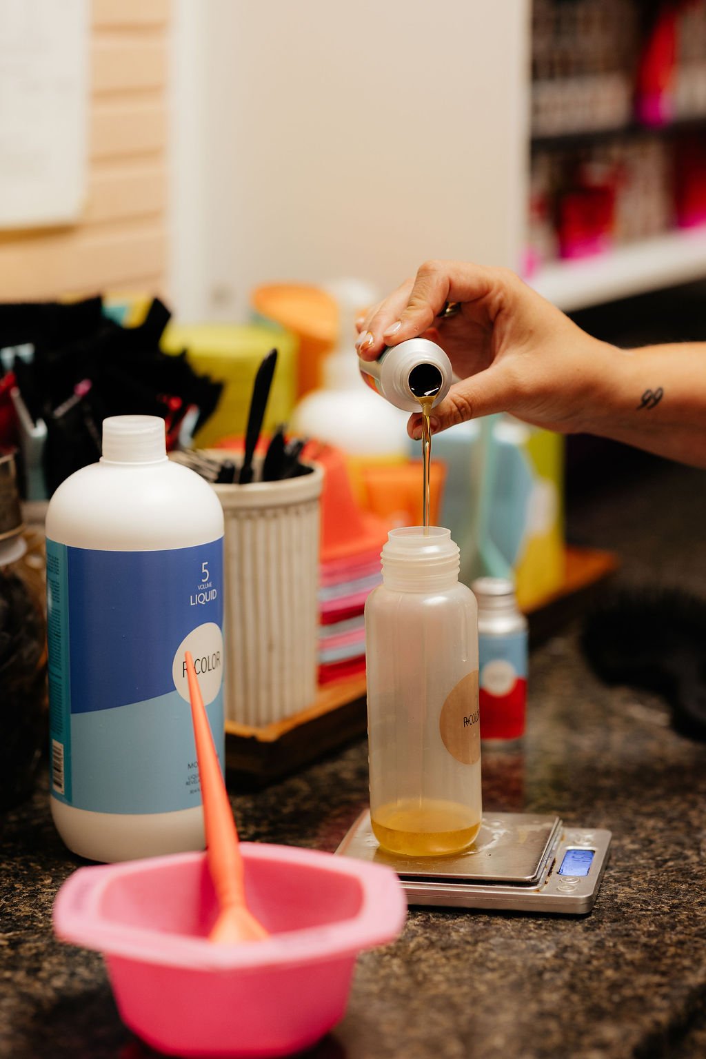 A person is pouring a liquid into a plastic bottle on a digital scale in a workspace, with various bottles and containers in the background.