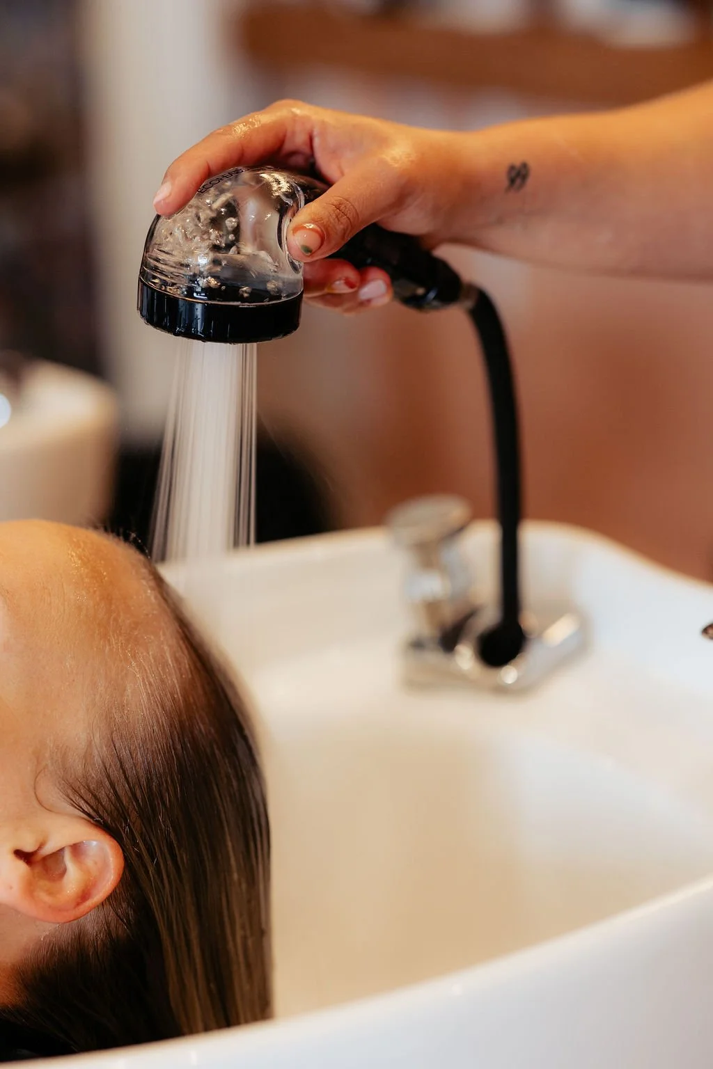 A person is washing a person's hair in a salon sink with a handheld showerhead.