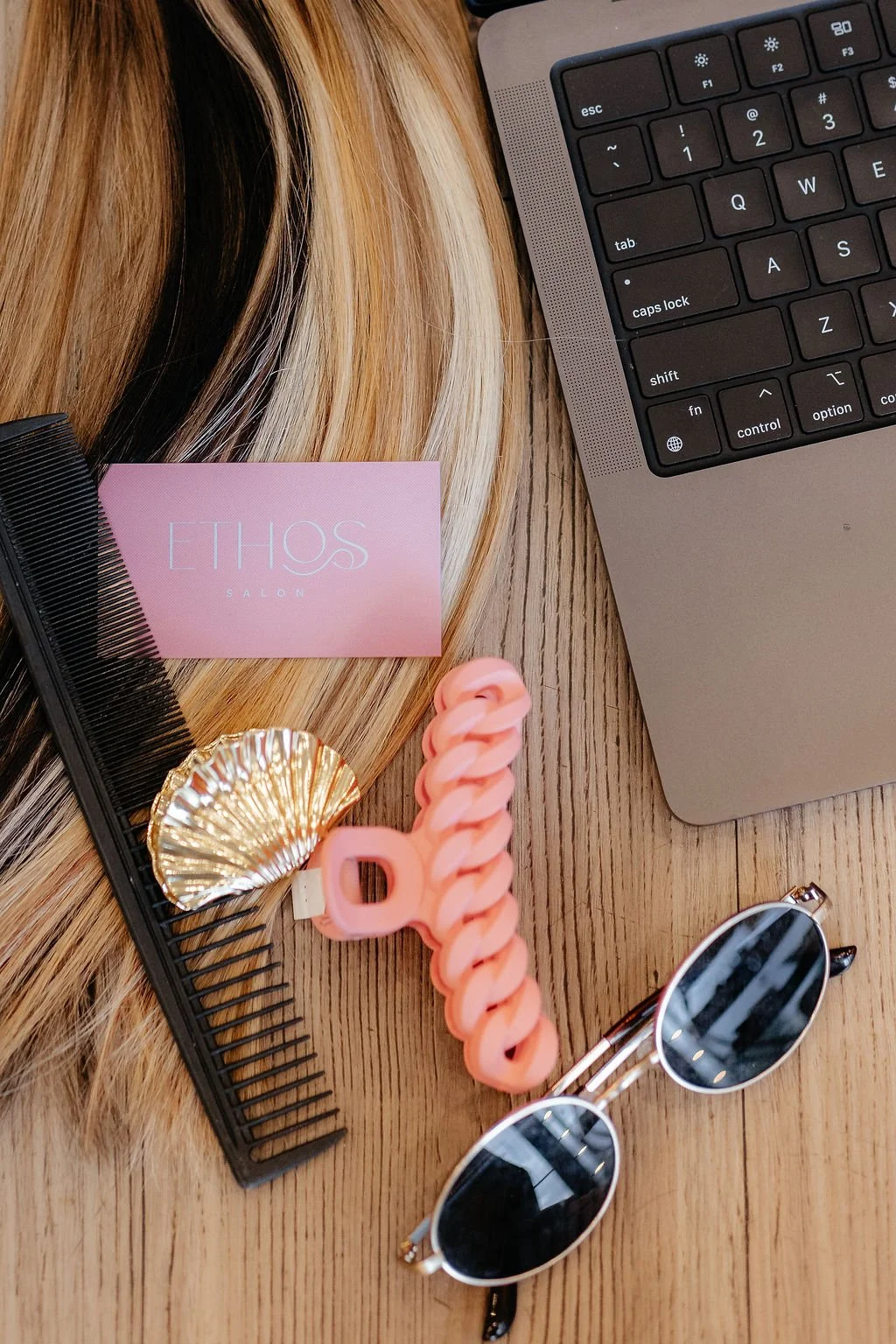 A flat lay of a wooden table with blonde hair, a pink card reading "ETHOS SALON," a black hairbrush, a gold decorative seashell, pink hair clips, a pair of sunglasses with black lenses, and a laptop keyboard.