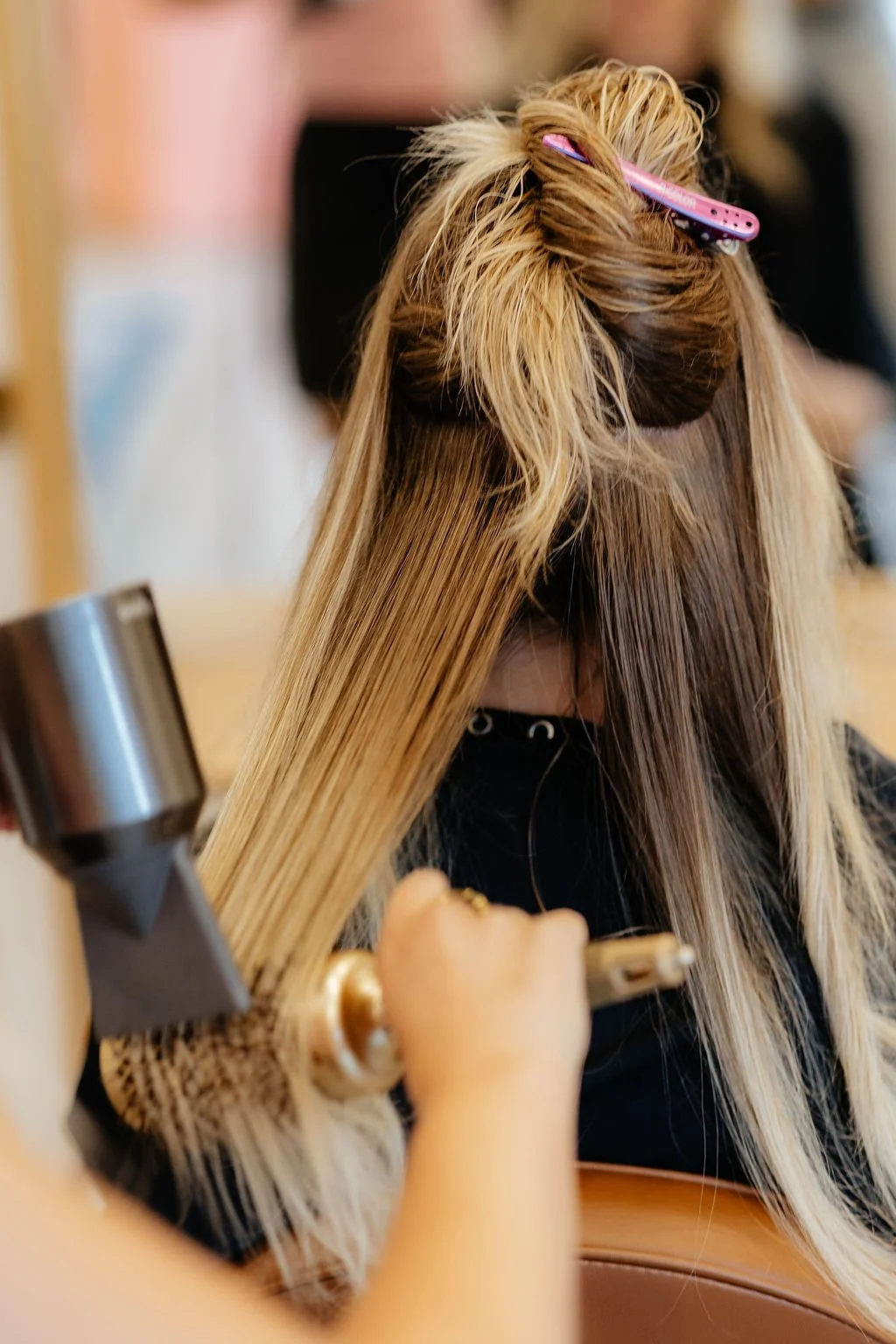 A woman with long, blonde hair getting her hair styled, with a pink hair clip holding a section of hair up, while the stylist blows warm air on her hair.