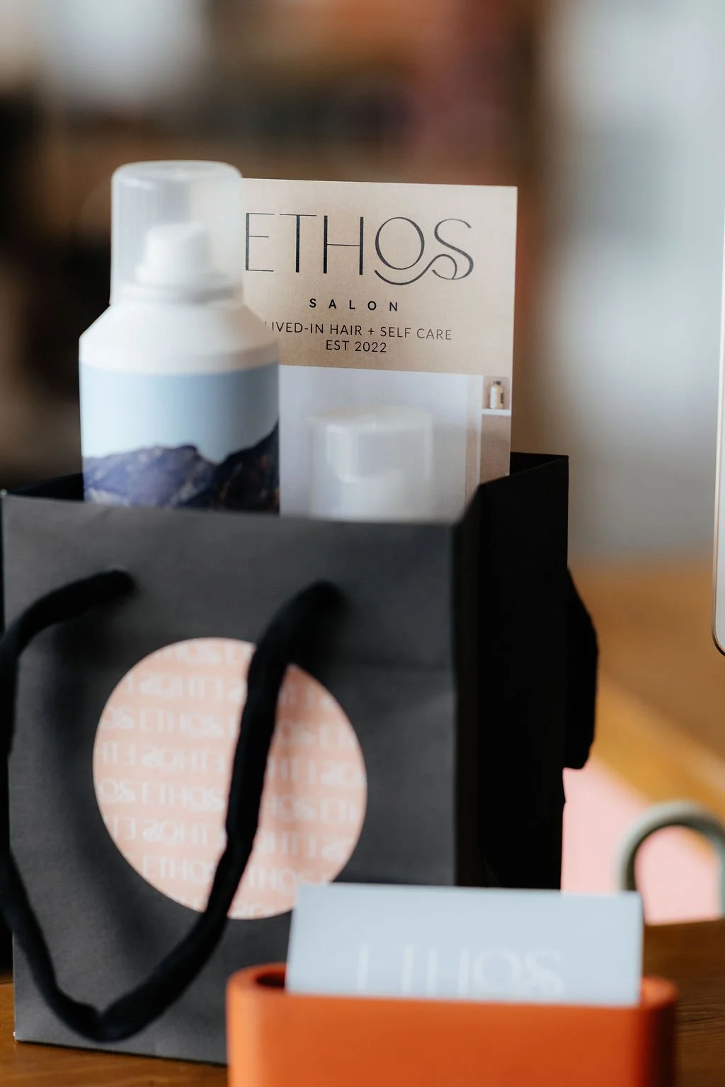 A black shopping bag with a peach-colored circle and black handles, a white bottle with a mountain landscape on the label, and a brochure for ETHOS Salon inside the bag.