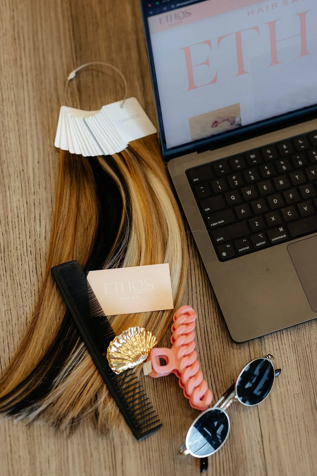 A wooden table with a laptop, a hair color swatch, a hair extension, a black comb with a white label, a gold decorative shell, a pink hair clip, and a pair of round sunglasses.