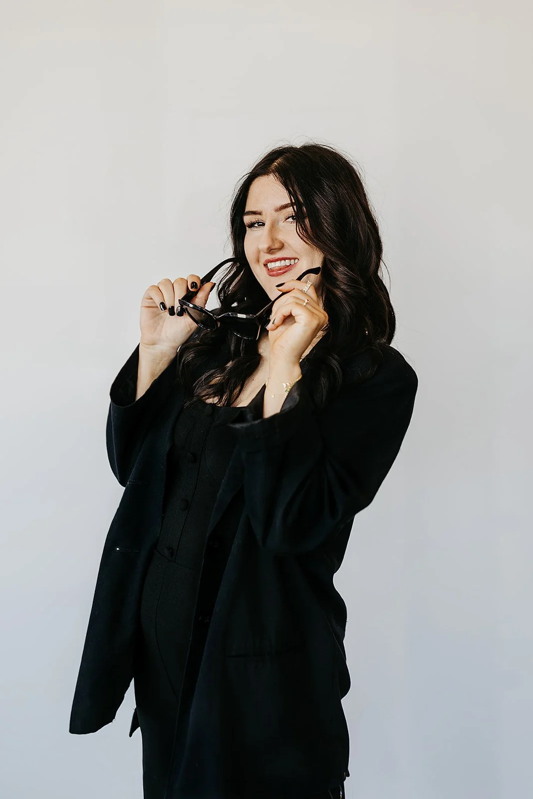 Woman with dark wavy hair wearing black outfit, smiling, holding black sunglasses.
