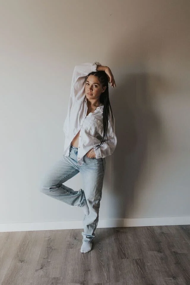 A young woman standing against a plain wall, wearing a white shirt and light blue jeans, with one leg bent and her hand resting on her head.