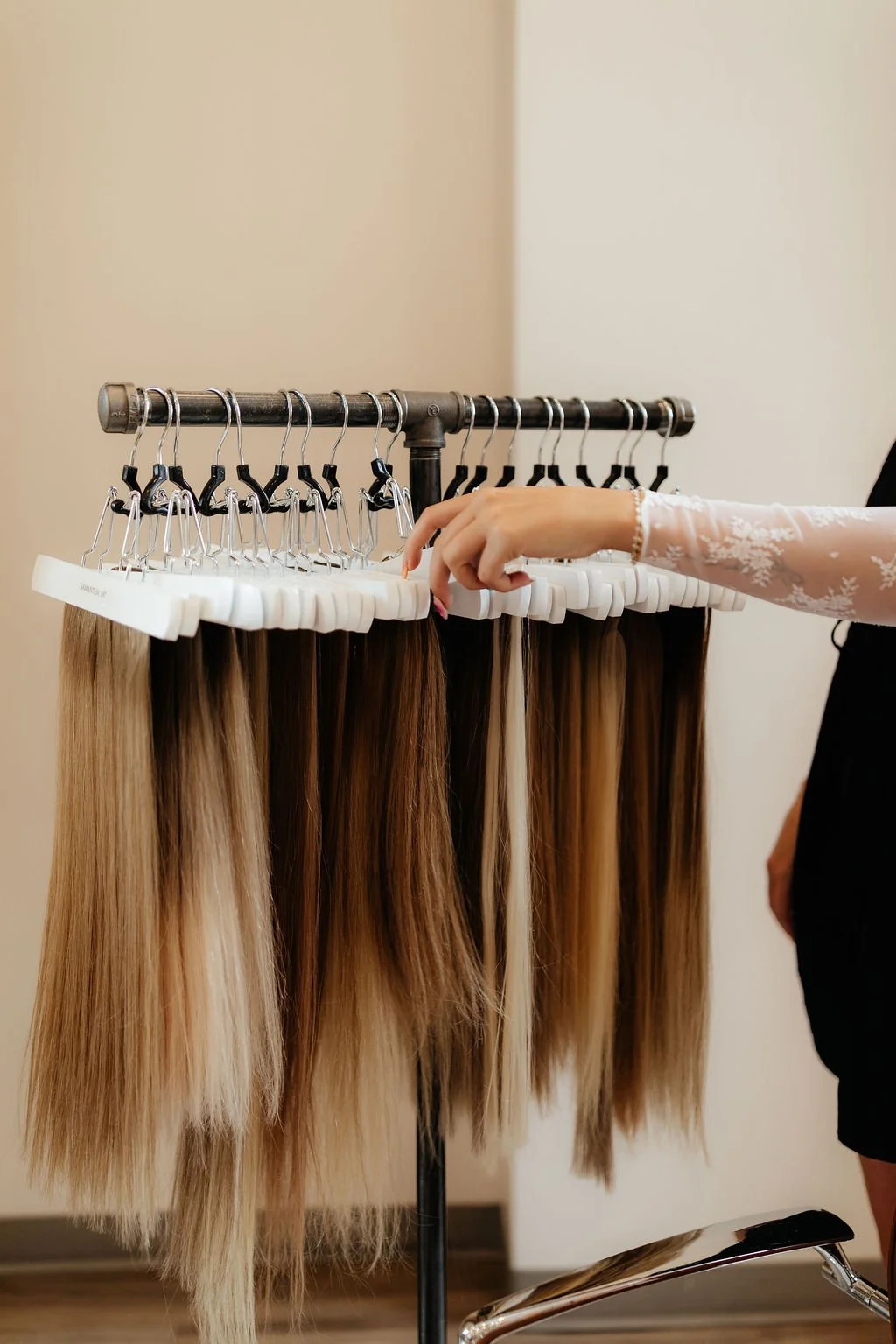 A person with a lacy white sleeve selecting a hair extension from a rack of blonde and brown hairpieces at a salon.