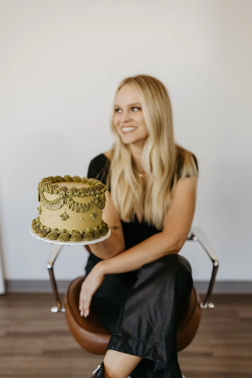 A woman with long blonde hair and a black dress sitting on a brown chair, holding a decorated cake with gold details, in front of a plain white wall.