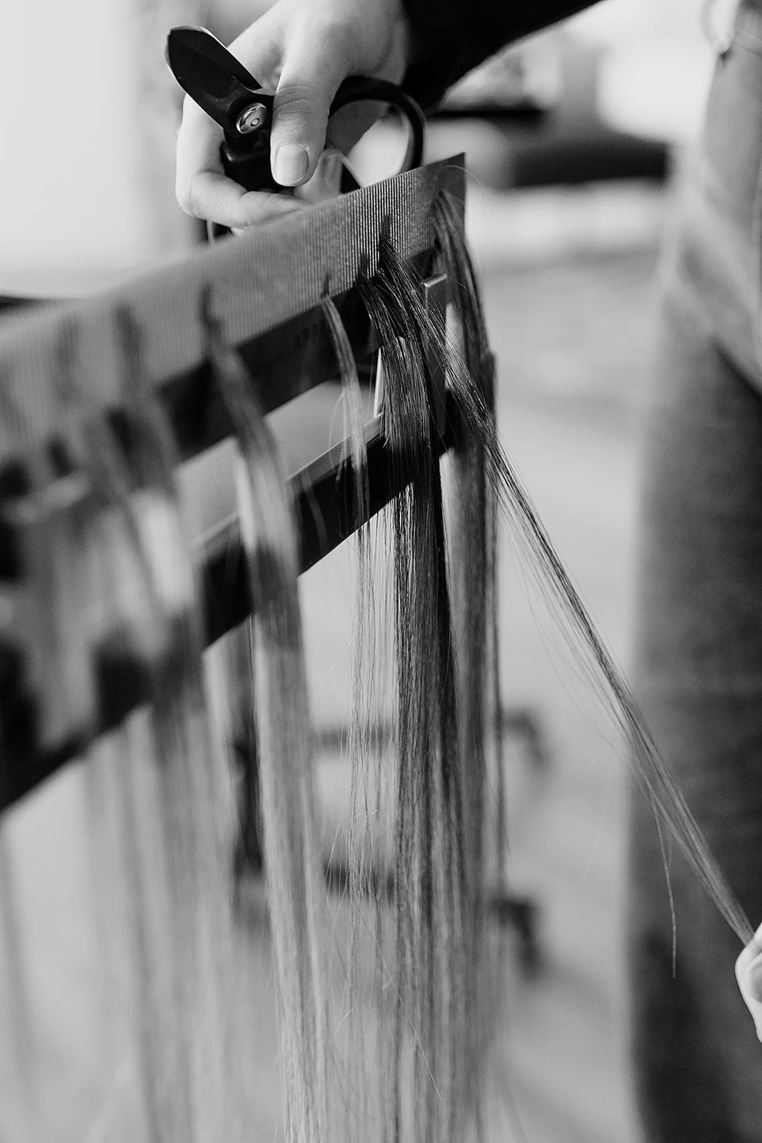 Black and white photo of a person's hand holding a tool, possibly a spray bottle, positioned over a rack with long strands of hair or fiber hanging from it.