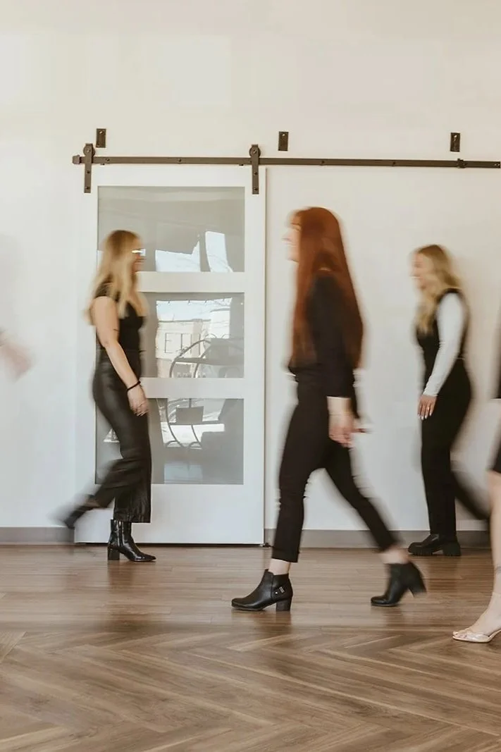 Three women walking past a plain white wall with a sliding barn door, hardwood floor, and blurred reflections on the door.