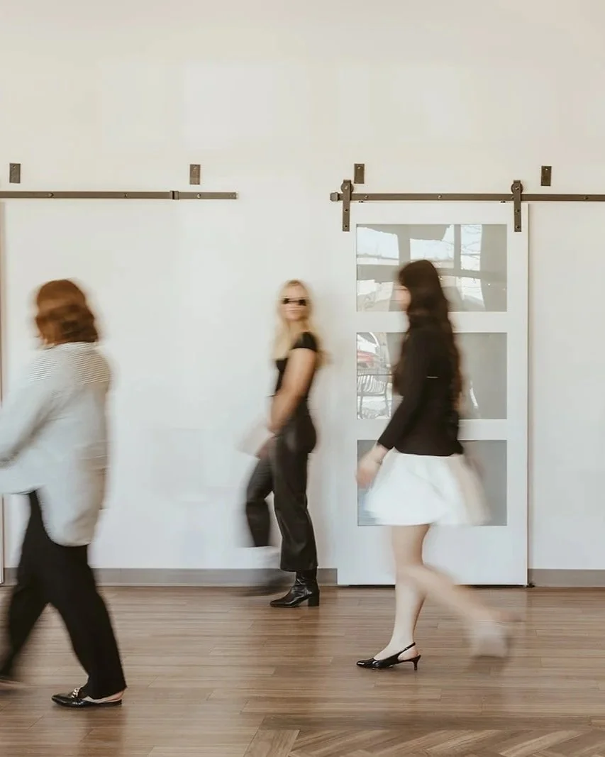 Three women walking in front of a white sliding barn door with glass panels, with one woman leaning against the door, all dressed in fashionable clothes.