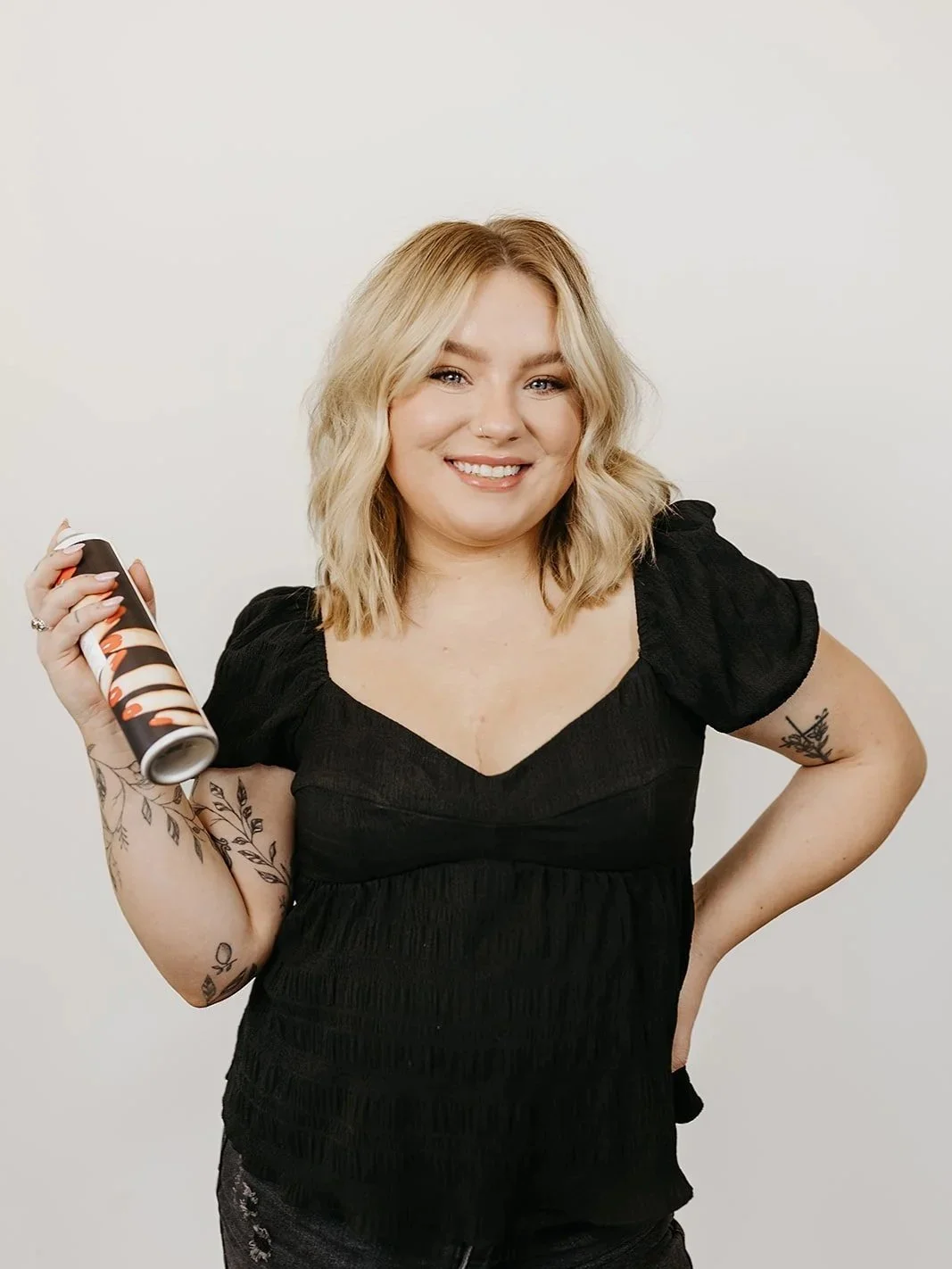 A young woman with blonde, wavy hair smiling while holding a can of hairspray in her right hand, wearing a black top with puffed sleeves, tattoos on her left arm, and standing against a plain white background.