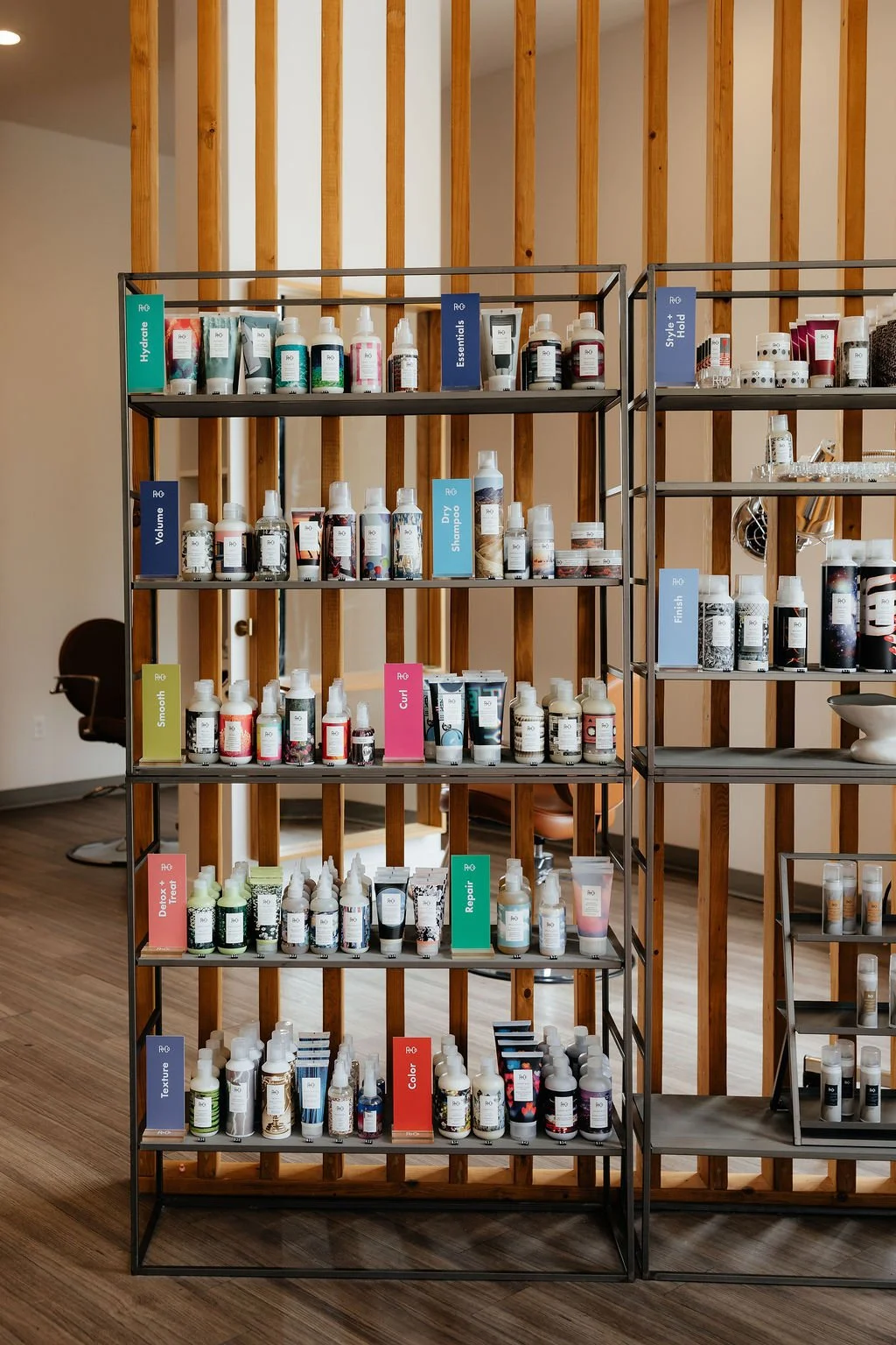 Display shelves in a salon with various hair care products organized by categories such as hydrate, essentials, style & hold, volume, dry shampoo, finish, smooth, curl, detox & treat, repair, texture, and color.