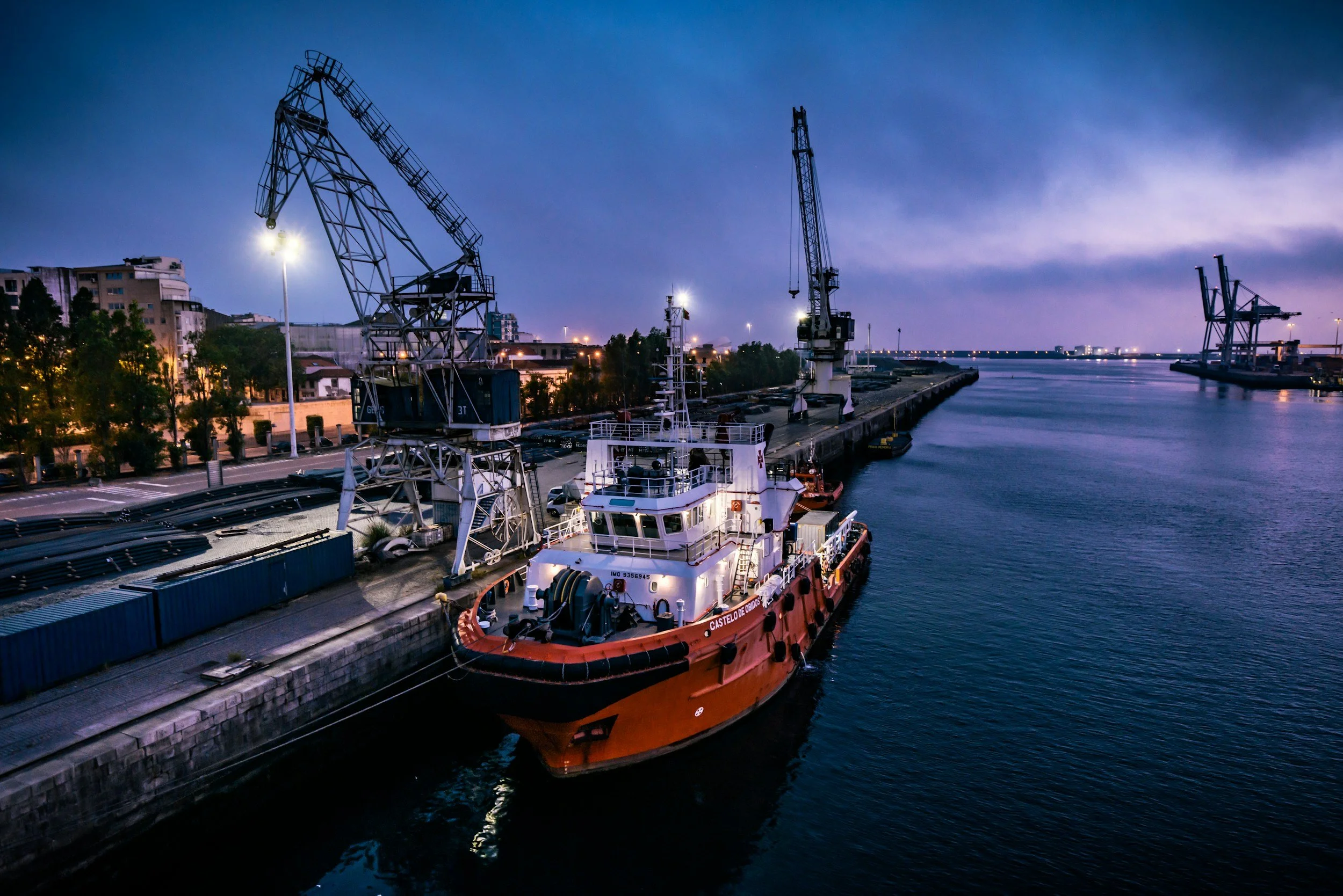 A harbor at dusk with a small orange and white tugboat docked along the pier, cranes, and industrial buildings in the background under a darkening sky.