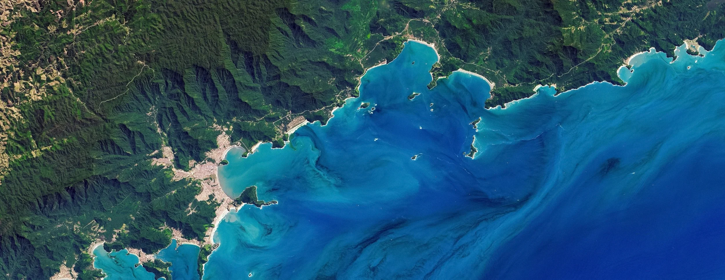 Satellite view of a coastal area with mountains and several bays filled with turquoise and blue water, and a developed shoreline with towns and roads.