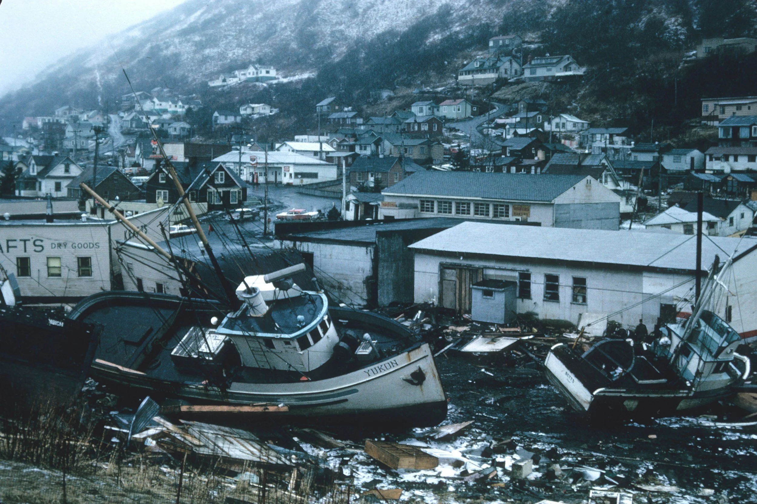 Damaged boats and debris in a flooded harbor with buildings and houses in a mountainous area in the background.