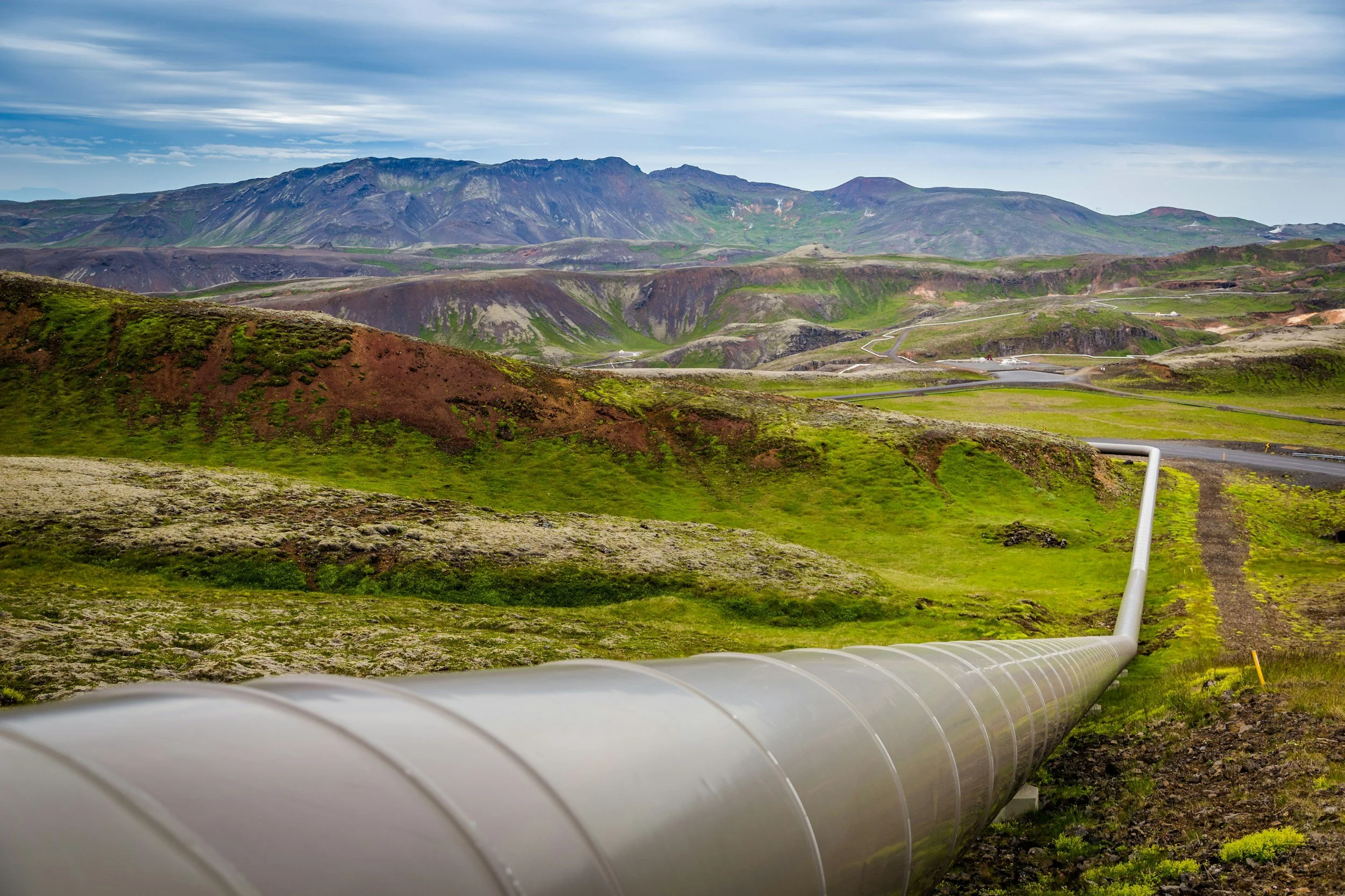 A large pipeline running across a lush, green landscape with hills and mountains in the background, under a partly cloudy sky.