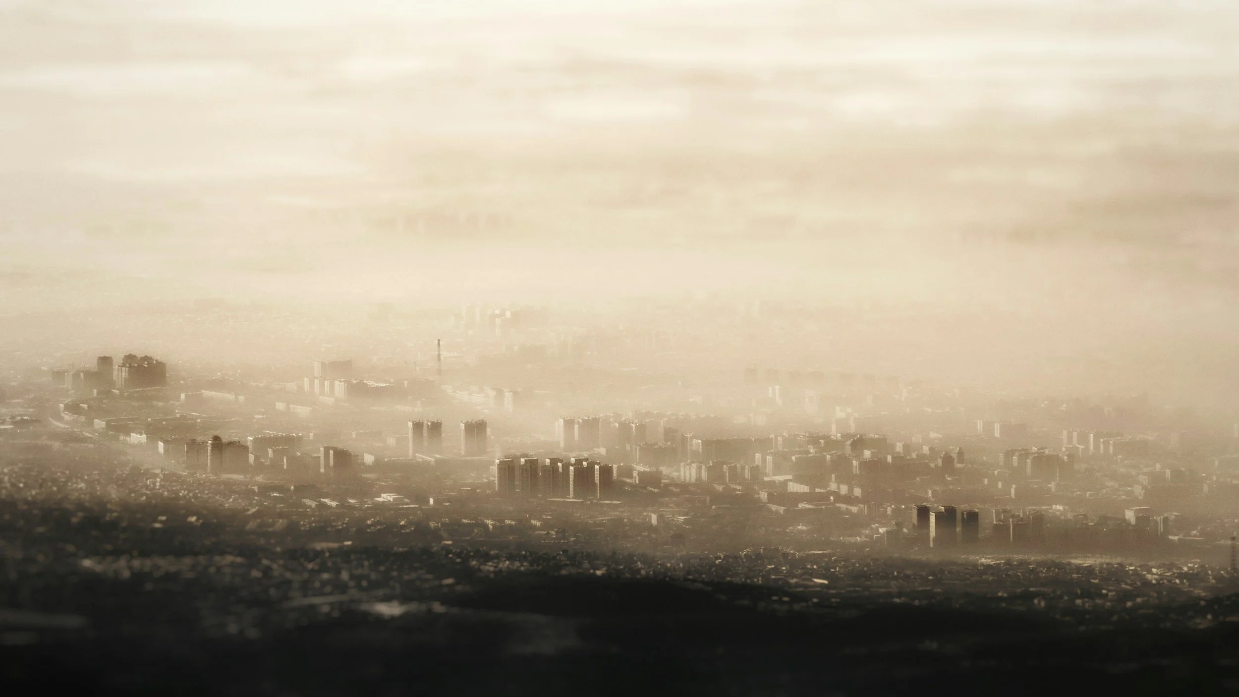 A cityscape with tall buildings and skyscrapers under a hazy, overcast sky, viewed from a distance.