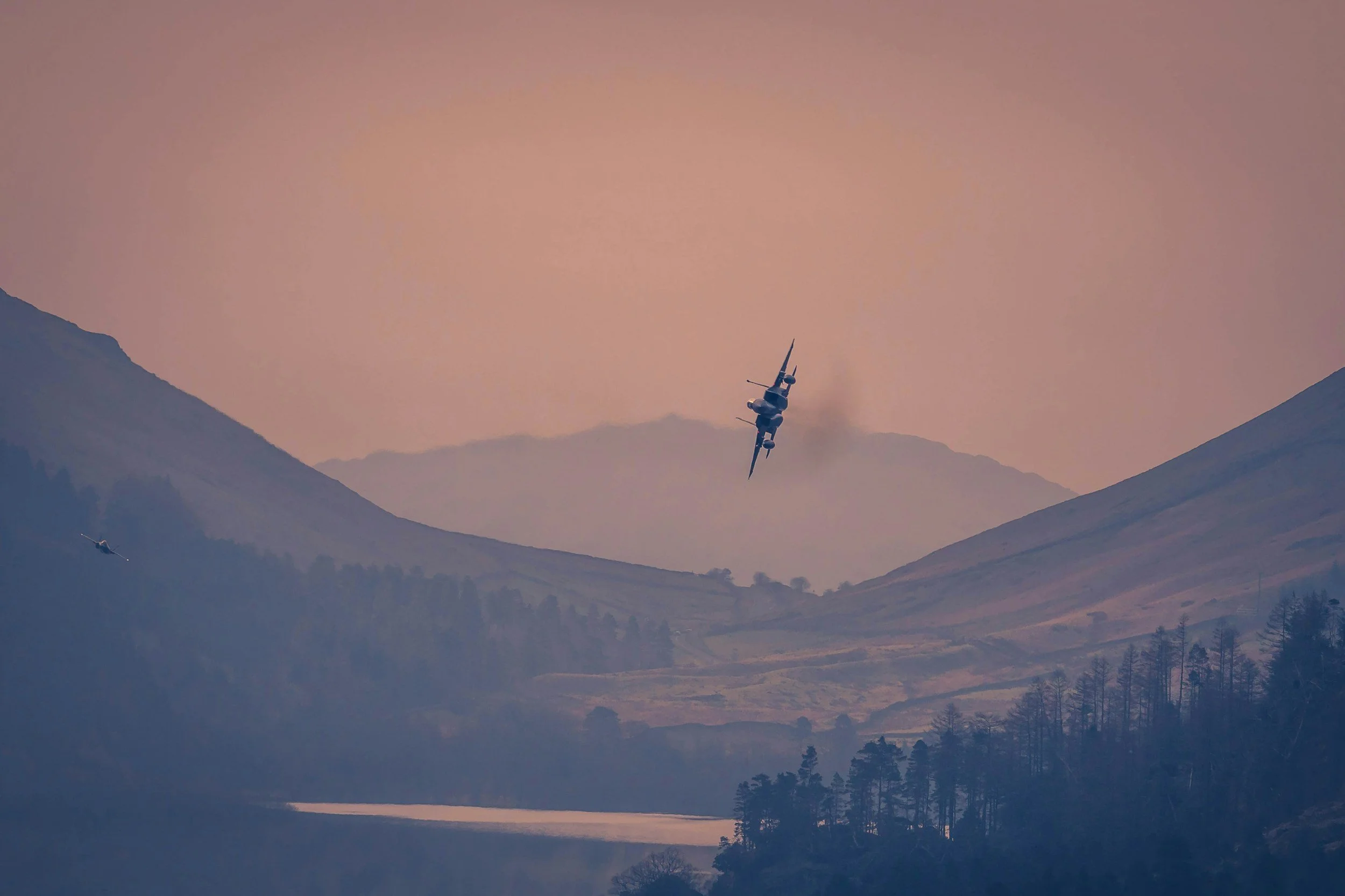 A fighter jet flying at an angle over a mountainous landscape during sunset or dusk.