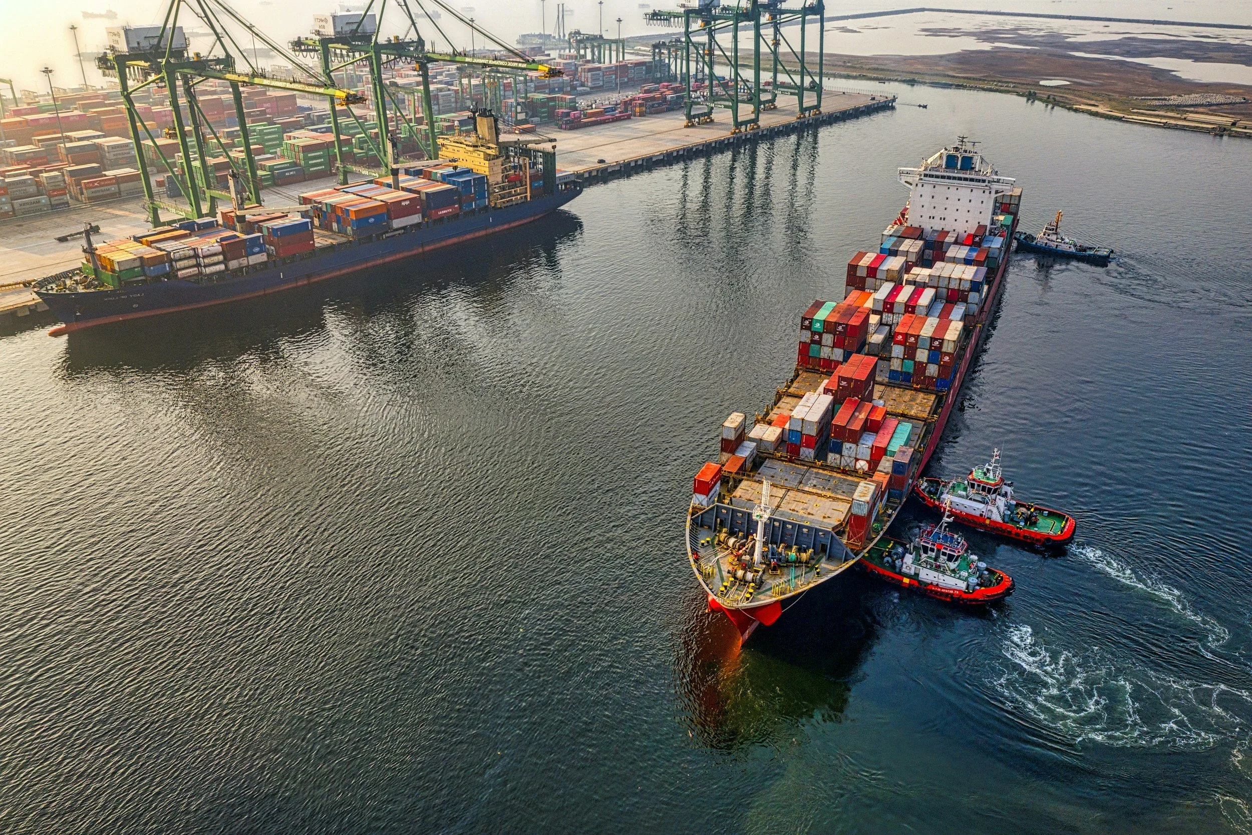 An aerial view of a shipping port with a large cargo ship and containers being loaded or unloaded using cranes, with several tugboats assisting the vessel.