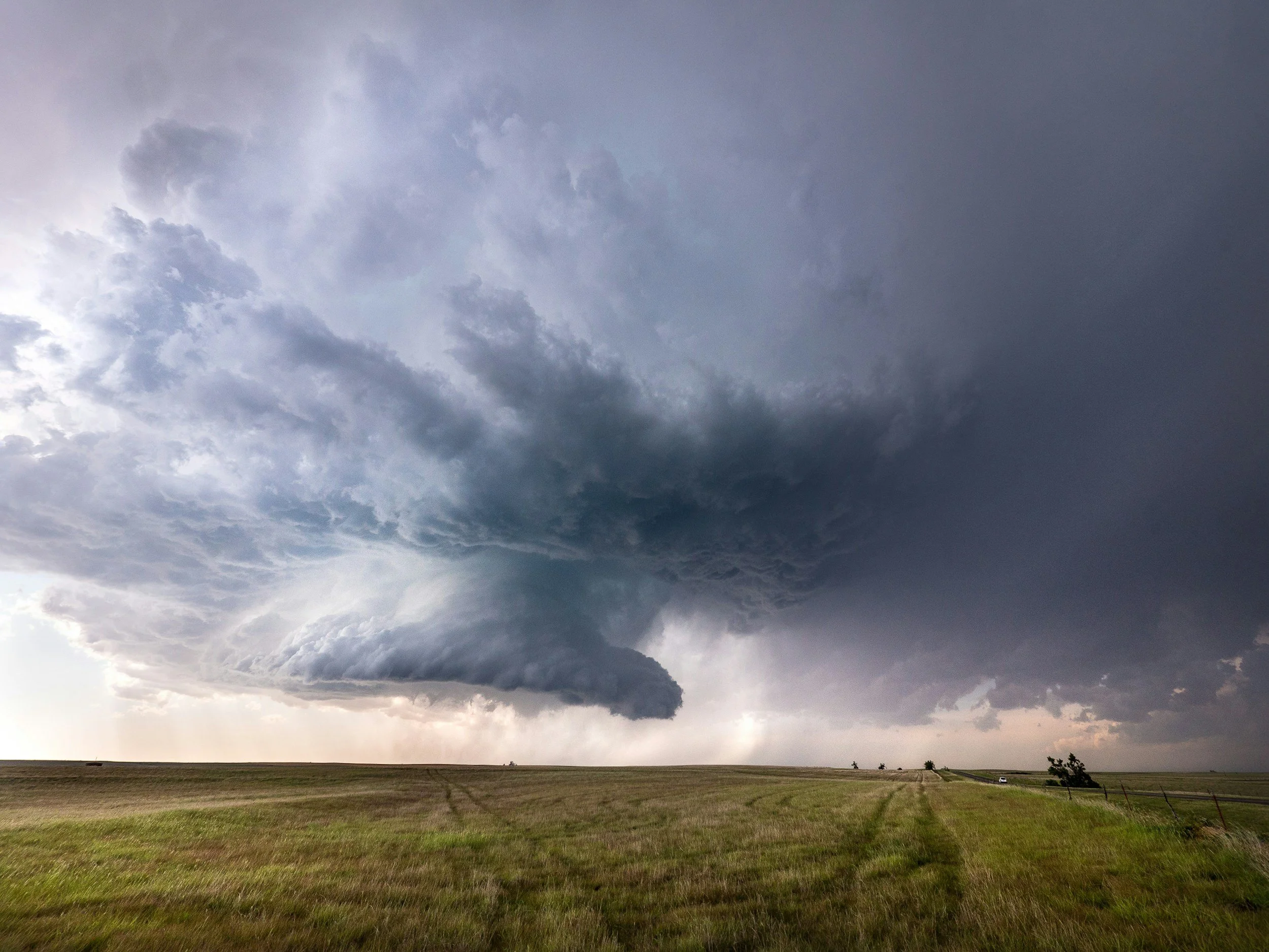 A large, dark storm cloud forming over an open grassy field with a few distant trees and a car in the distance.