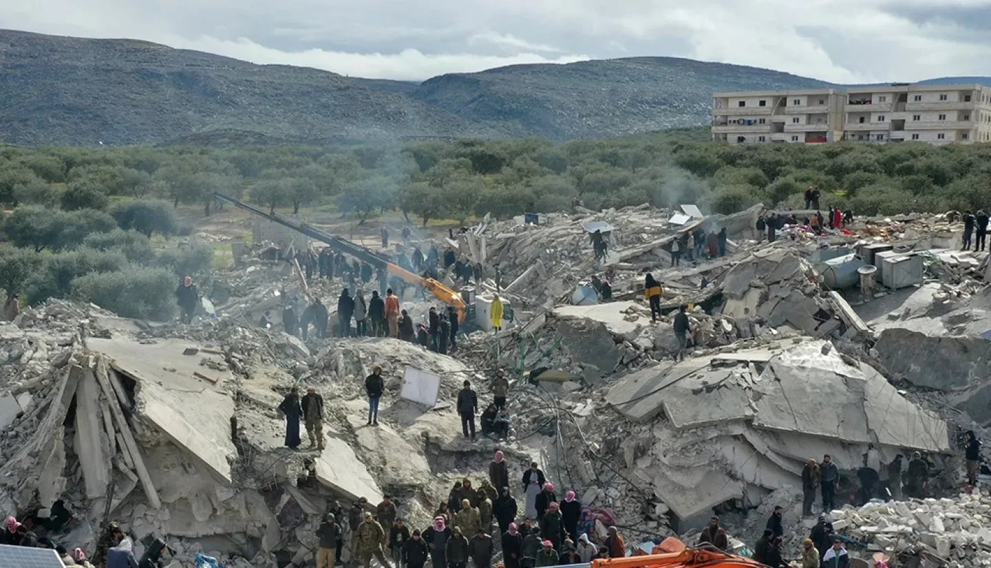 People at a collapsed building site with rubble and debris, in a mountainous area with nearby trees and an apartment building in the background.