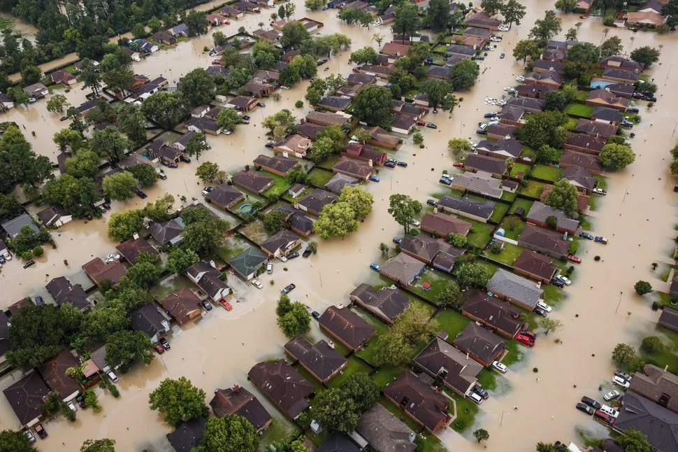 Aerial view of a neighborhood flooded with muddy water, with submerged houses, trees, and cars.