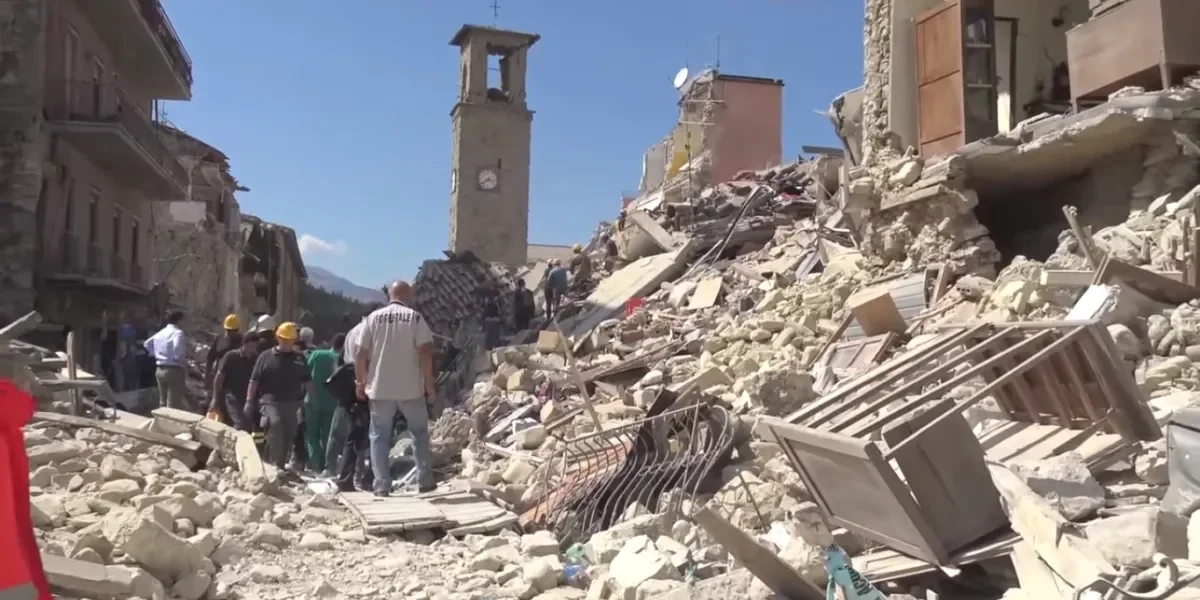 People walking among rubble and debris in an urban area with damaged buildings after an earthquake.