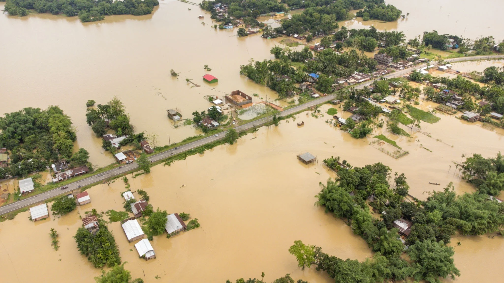 Aerial view of a flooded area with water submerging houses, trees, and roads. Floodwaters are muddy brown, with some structures partially underwater and debris floating.