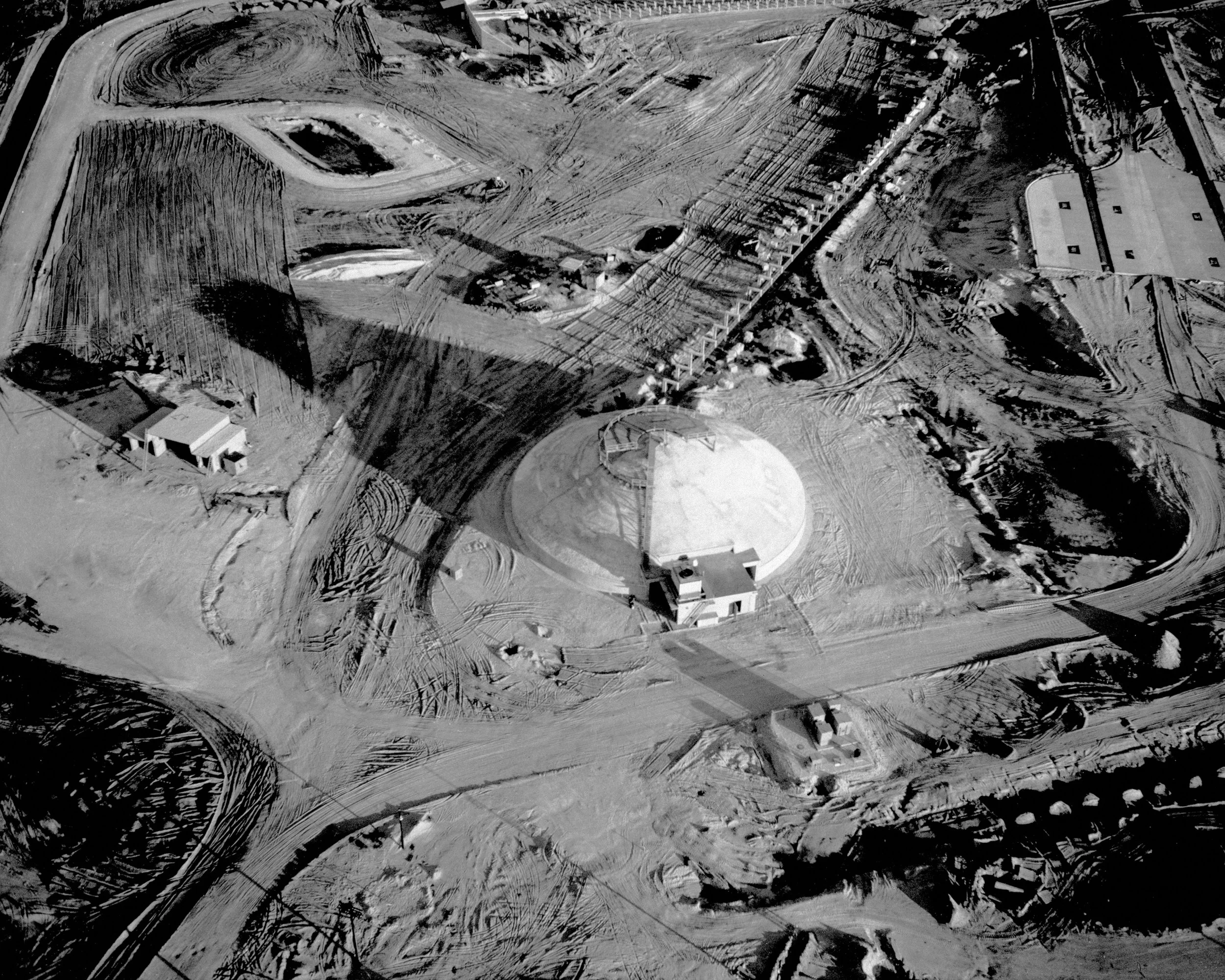 Aerial view of a water treatment plant under construction, with large circular tanks, buildings, and numerous pathways and dirt roads on a barren landscape.