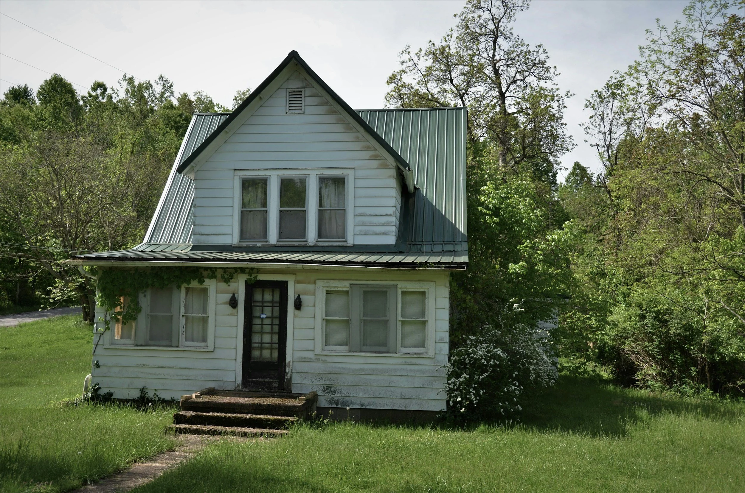 A two-story, white, wooden house with a green metal roof, surrounded by greenery and trees, with a small front porch and steps leading to the front door.