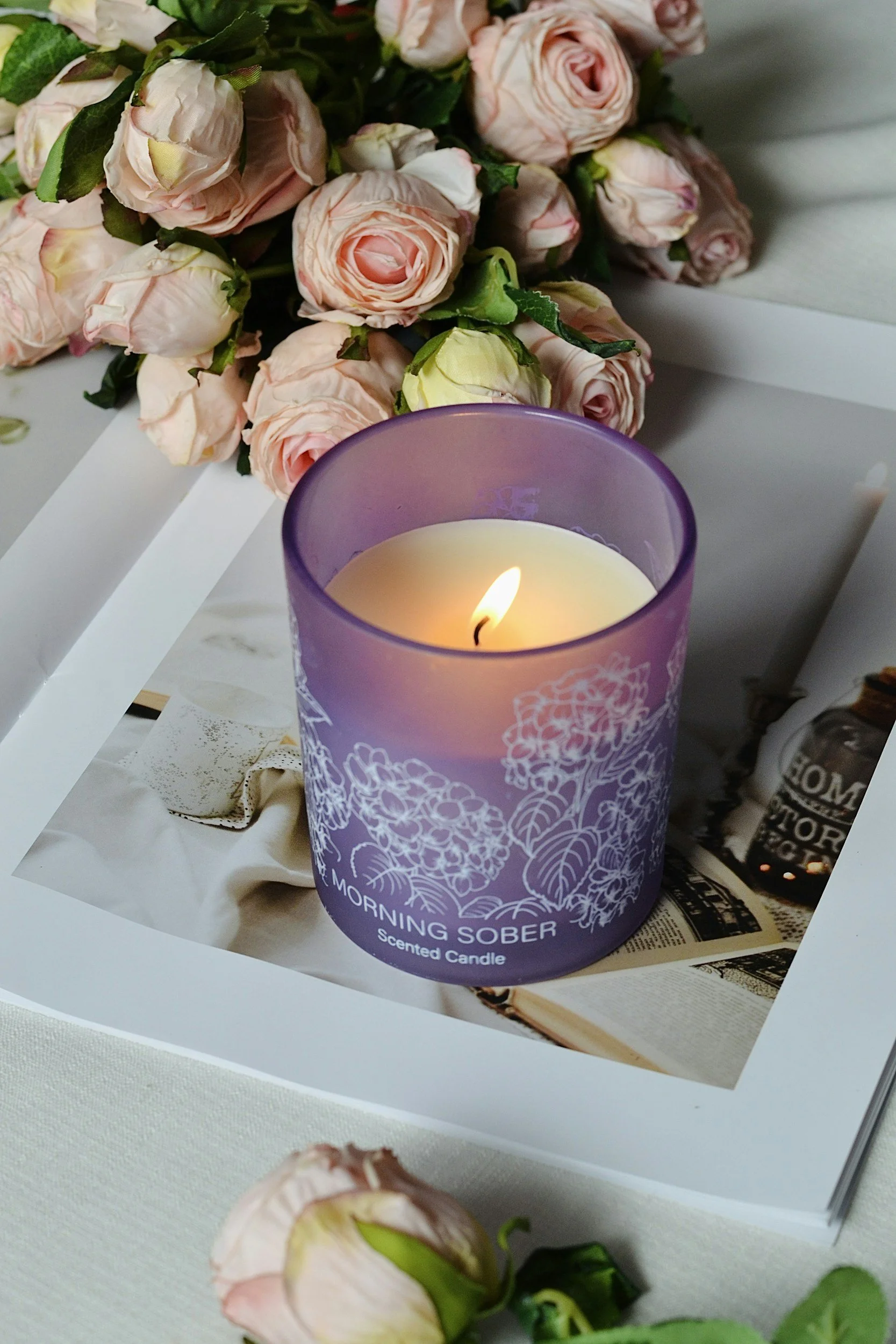 A lit scented candle labeled 'Morning Joy' in a purple glass container, with a bouquet of pink roses in the background, and an open book and other items on a desk.