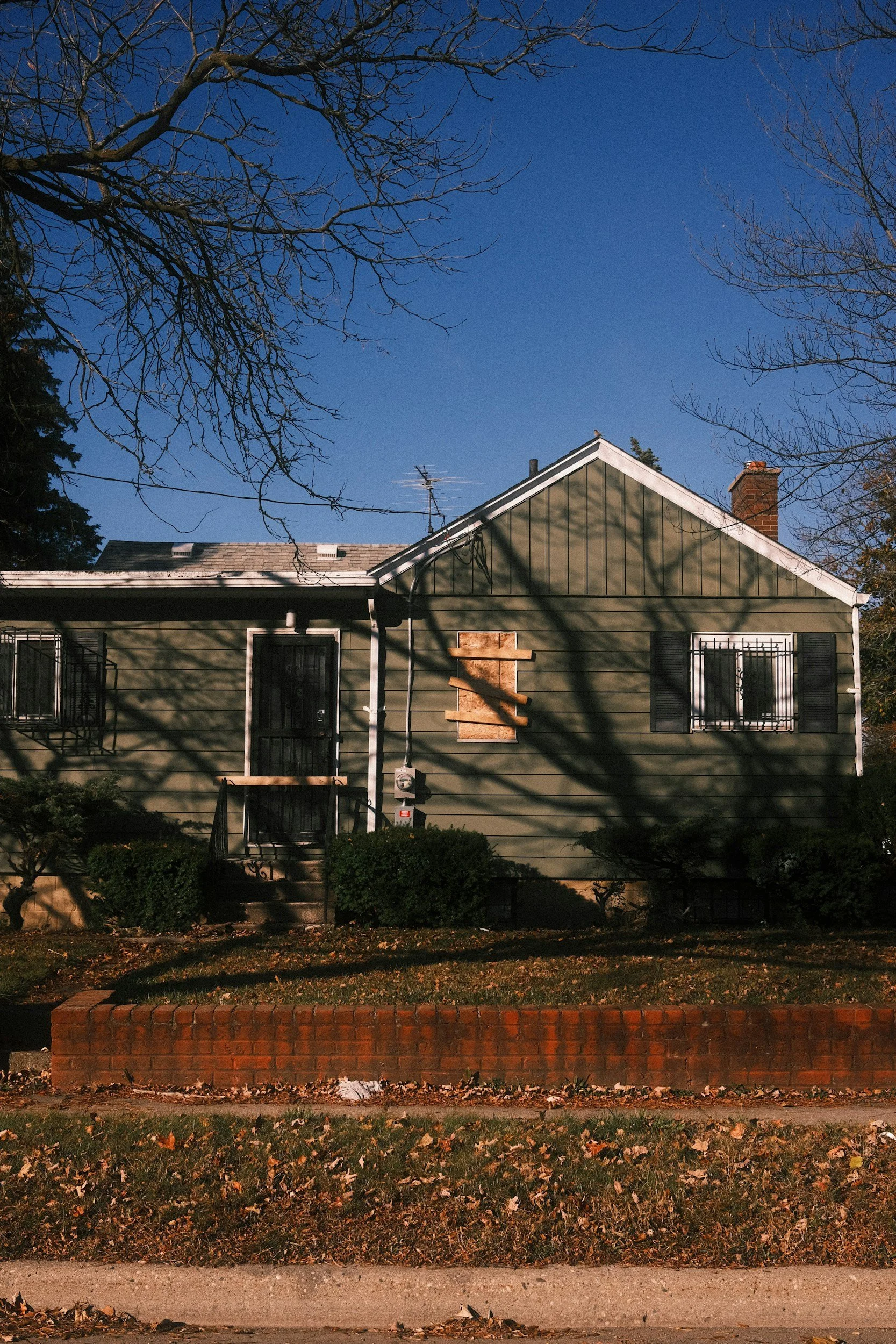A house with green siding and boarded-up window, shadows of tree branches on the facade, in a neighborhood with leaves on the ground and bare trees.