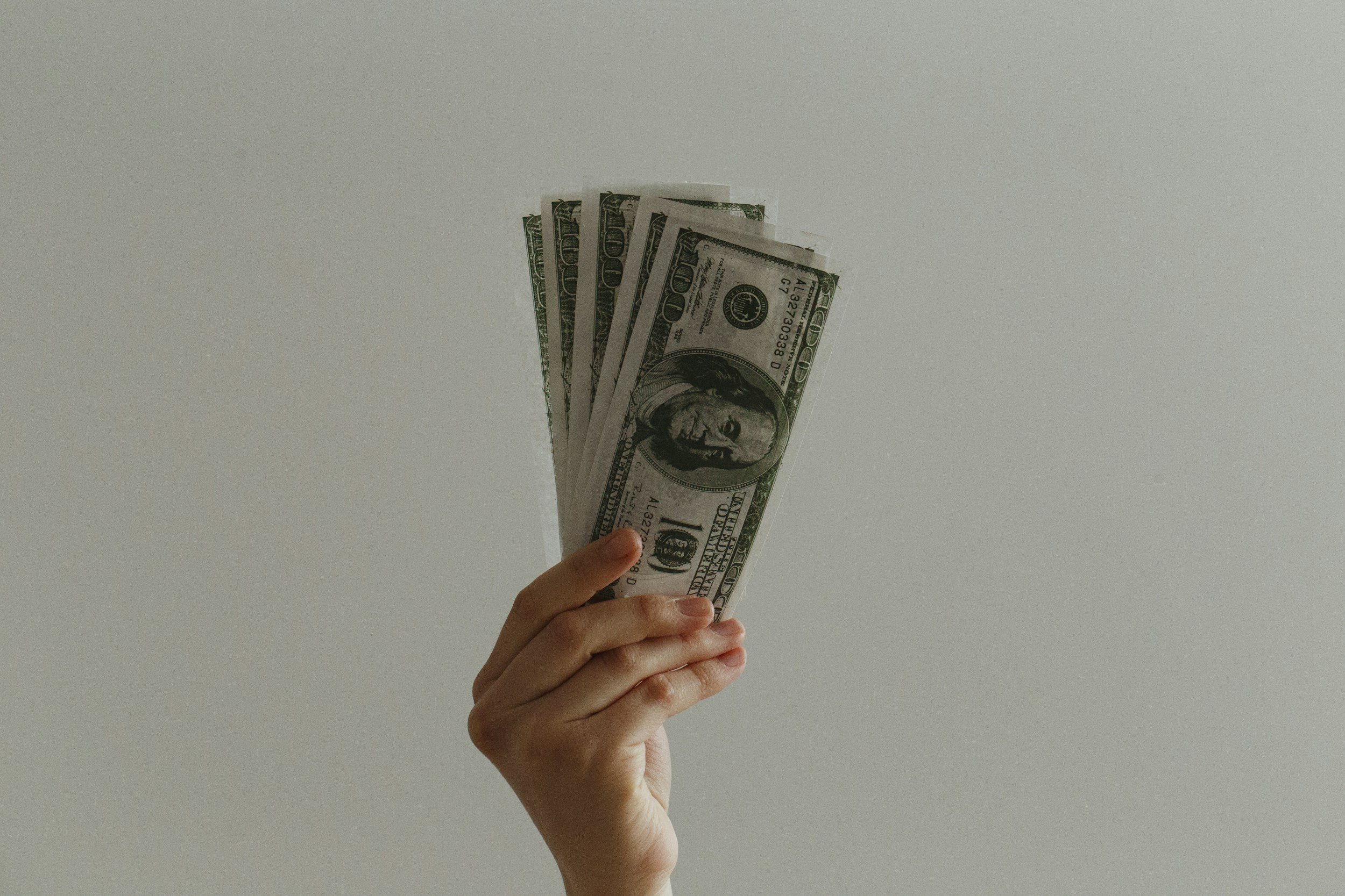 Hand holding a fan of US hundred-dollar bills against a plain white background.