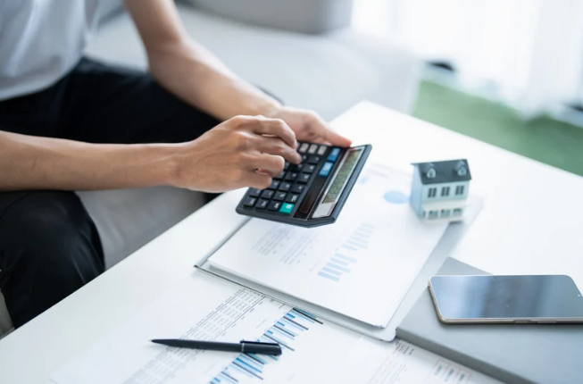 Person using a calculator on a desk with documents, a pen, a tablet, and a small house model.