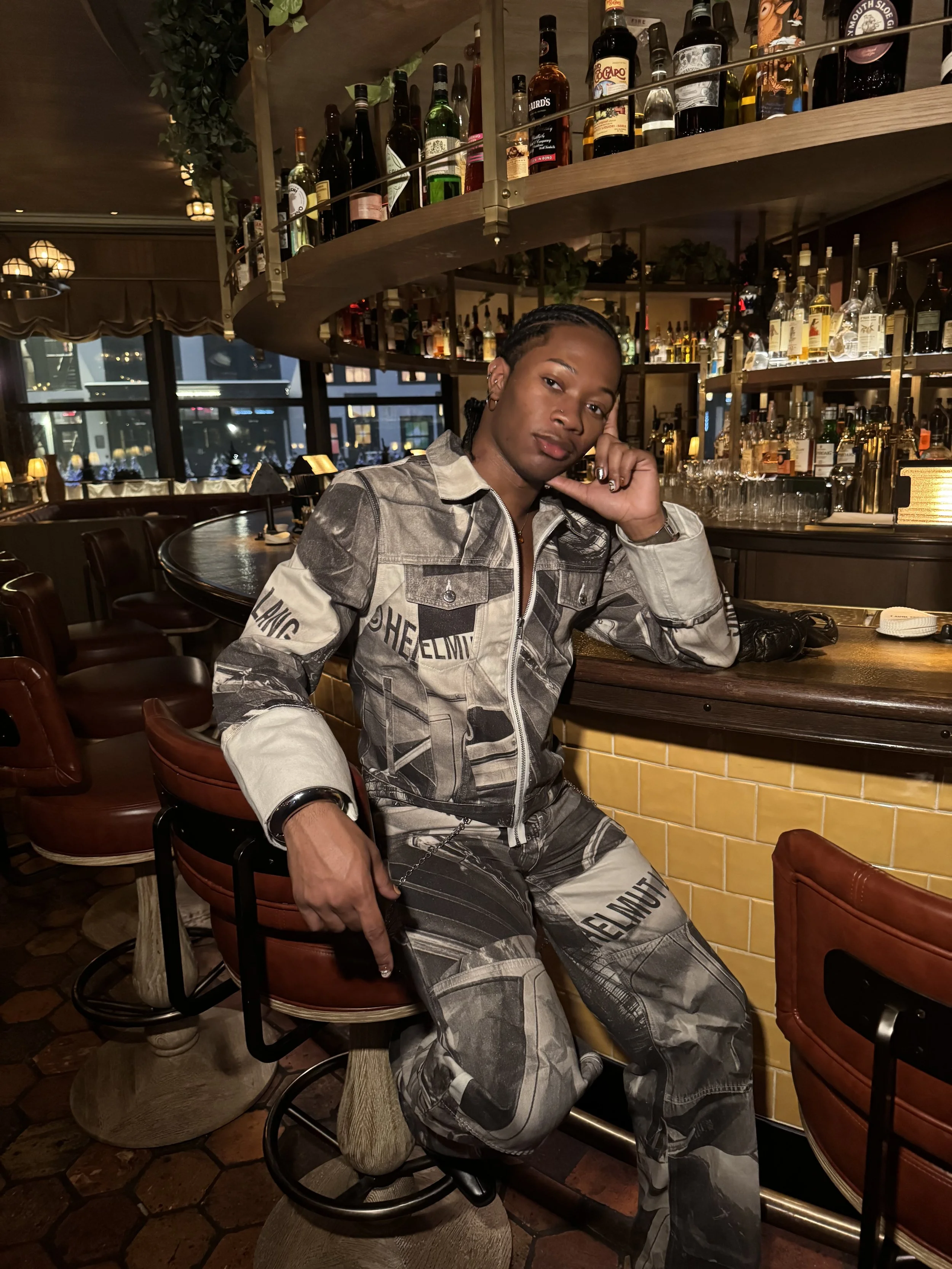 Waverly sitting at a bar in a dimly lit restaurant or bar, with shelves of liquor bottles behind them, a window showing city lights outside, and warm ambiance lighting.