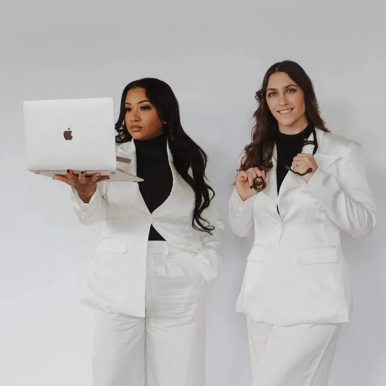 Two women dressed in white medical coats, one holding a laptop and the other with a stethoscope, posing against a plain white background.