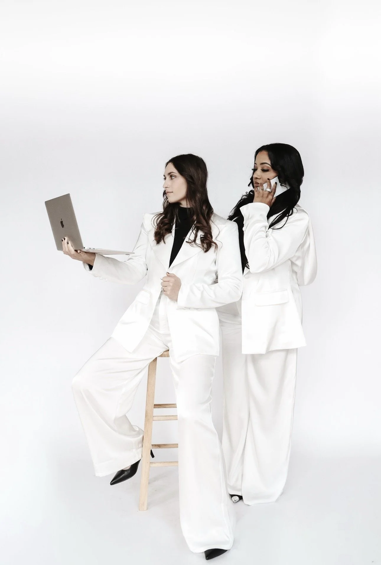 Two women dressed in white suits, one sitting on a stool with a laptop, and the other talking on a phone, standing against a white background.