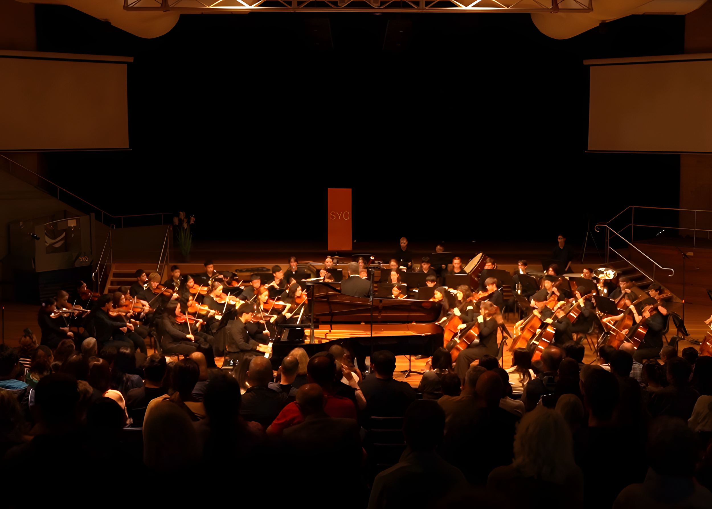 An orchestra performing on stage with a conductor in front, audience seated in front, large projection screens, and stage decorations including a sign with 'SYO'.